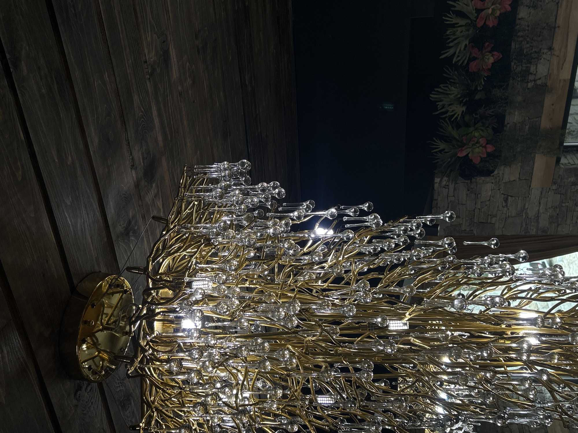 A close-up, low-angle shot shows a gold chandelier with clear teardrop-shaped glass accents hanging from a dark wooden plank ceiling. The chandelier is densely packed with thin, interwoven gold branches and numerous glass drops, some reflecting light from the bulbs within. Below the chandelier, a dark void is visible, with the upper portion of a stone wall and decorative plants with pink flowers partially illuminated. The image appears to be taken indoors, possibly in a restaurant or event space, in Batak, Bulgaria, though no specific location within the city is identifiable from the visual information alone.
