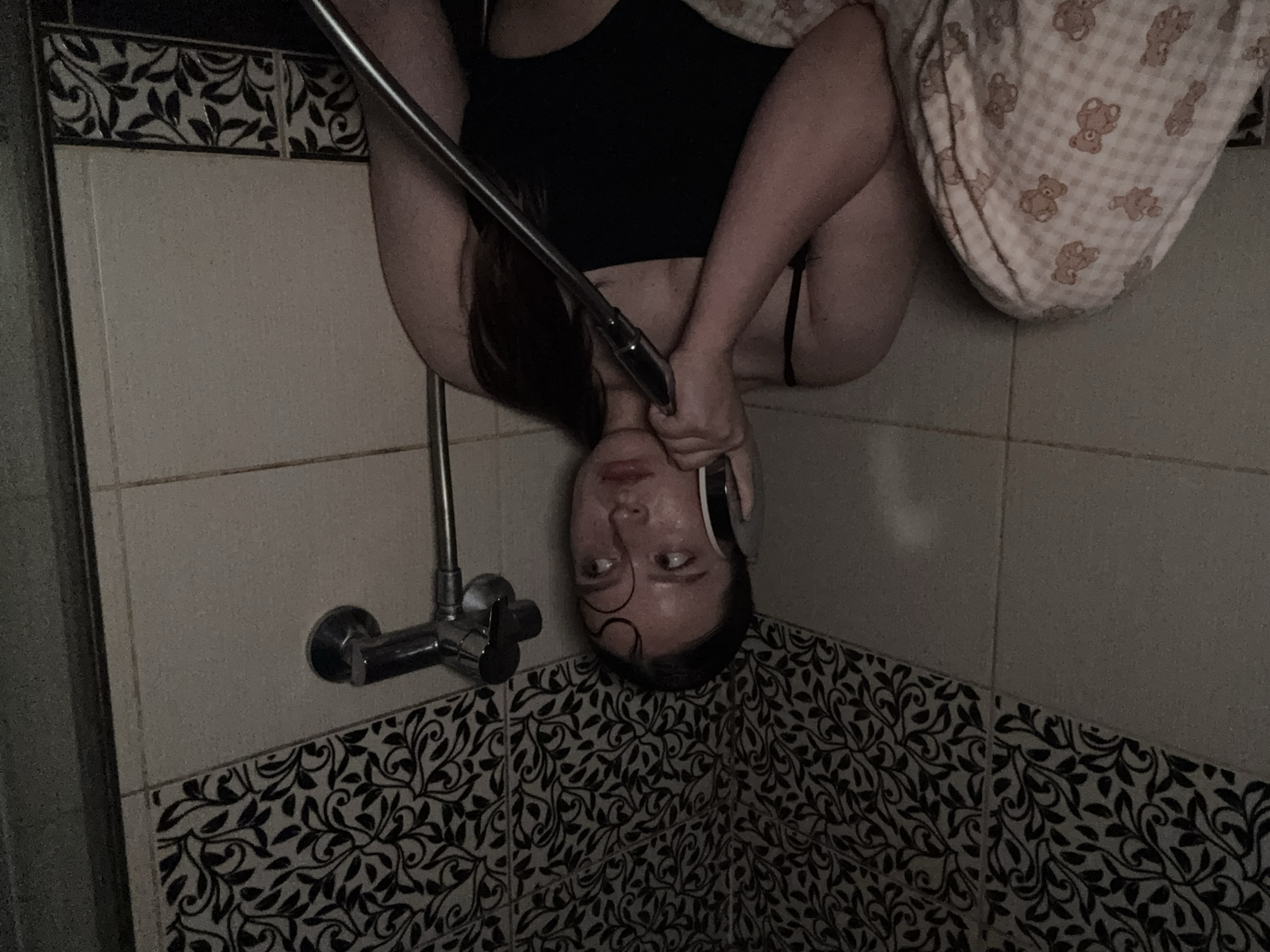 A female is depicted in a dimly lit bathroom environment. She has dark hair, which appears damp and is pulled back, with a few wet strands curled on her forehead. She is wearing a black spaghetti-strap top or swimsuit. Her lower body is partially covered by a light-colored blanket featuring a repeating pattern of small brown teddy bears on a checkered background. She holds a chrome-finished shower head to her left ear, as if using it as a telephone. Her gaze is directed to the right (her left), with wide eyes and a slightly parted mouth, conveying an expression of apprehension, concern, or surprise. The background consists of tiled walls; the lower section features plain light-colored rectangular tiles, while the upper section displays decorative tiles with a dark, intricate floral or leafy pattern on a lighter background. A chrome shower mixer unit is visible on the wall to her right. The overall lighting is low, contributing to a subdued atmosphere.