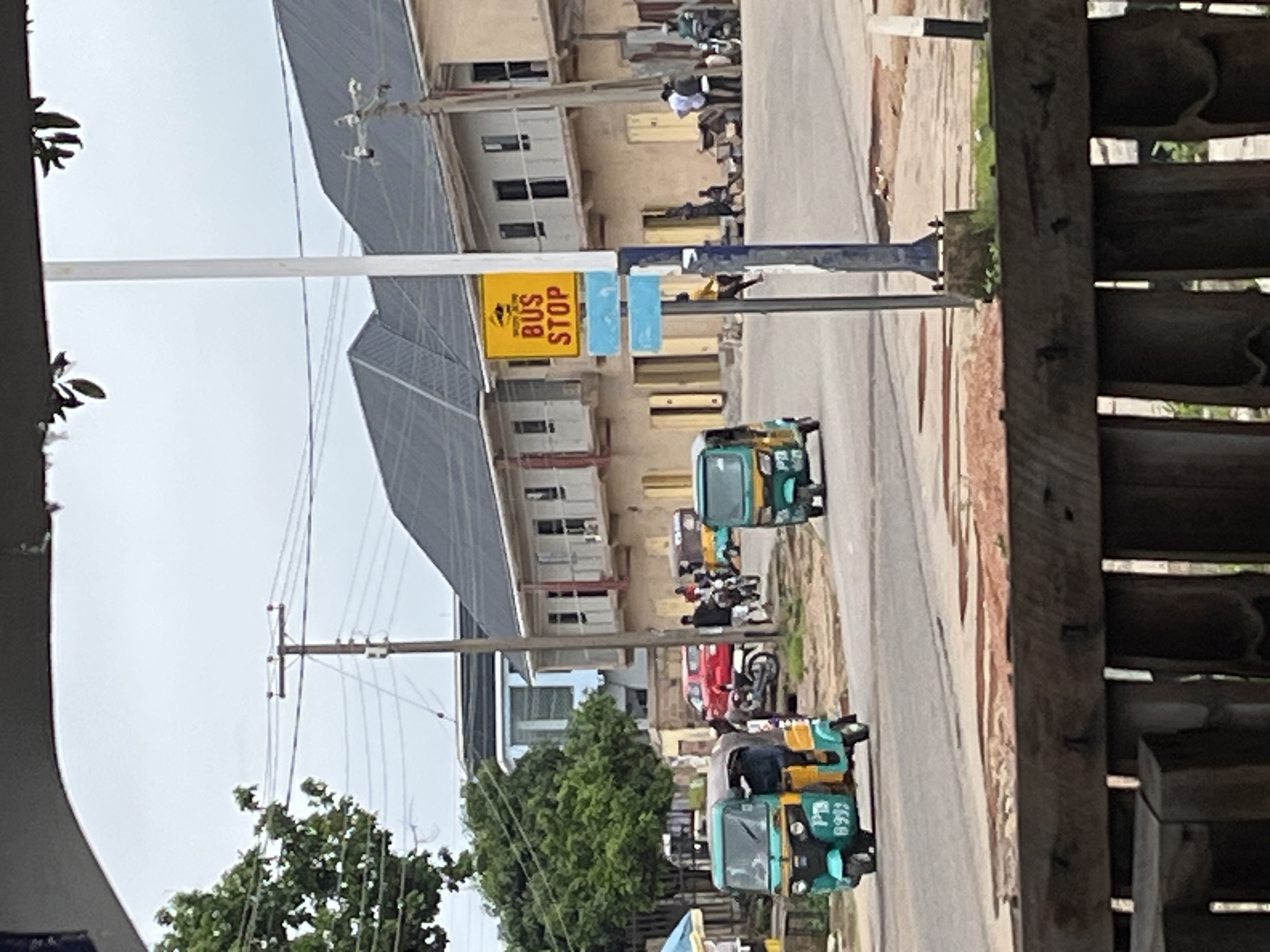 The image captures a street scene in Jos, Nigeria, likely during daytime, characterized by a mix of residential and commercial structures. A bus stop sign, prominently featuring "BUS STOP" in yellow with red lettering, is affixed to a pole on the right side of the road. Several auto-rickshaws, commonly known as Keke Napep, are visible, with two in the foreground and one further down the street. One of the Keke Napep in the foreground is green and yellow, with the license plate "PB 6993" clearly visible.
The road appears to be paved, with some reddish-brown patches suggesting a mixture of dirt and asphalt. Beyond the road, a multi-story building with a dark, sloped roof and numerous windows dominates the background. Adjacent to it is another building with a lighter facade and balconies. A red car is parked on the side of the road, and several individuals are seen standing or walking near the buildings and the road. Trees with green foliage are present on the left side of the image, and utility poles with wires crisscross the sky. In the very foreground, a blurred wooden structure, possibly a fence or bench, partially obstructs the view. The overall atmosphere suggests an active urban environment.