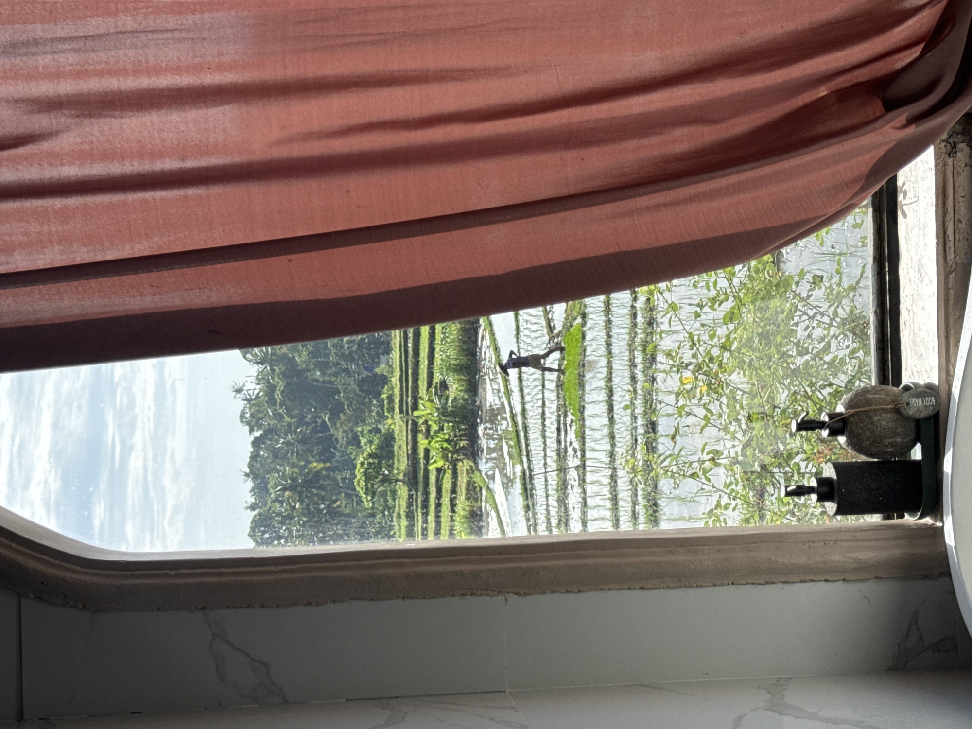 The image, captured in Mengwi, Indonesia, depicts a view through a window onto a lush landscape. In the foreground, a portion of a pink curtain is visible on the right side of the frame, and on the left, a marble-patterned wall and window frame. The windowsill holds two soap dispensers and a decorative stone. The window looks out onto tiered rice paddies, reflecting the sky and surrounded by dense green foliage and trees. A lone figure, possibly a farmer, can be seen in the middle ground of the rice paddies, actively working. The sky above is partly cloudy, with patches of blue.