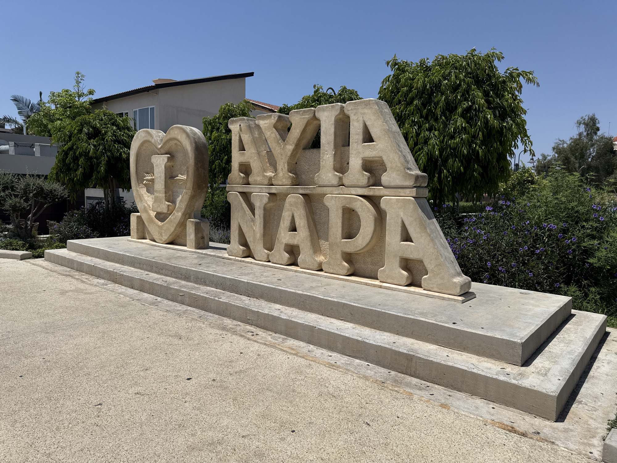 An outdoor sculpture featuring the words "AYIA NAPA" spelled out in large, stone letters on a concrete platform. The first part of the sculpture is a heart-shaped design with the letter "I" inside, also made of stone. The letters are monumental in size, carved from light-colored stone and have a rough, textured finish. The sculpture is situated on a paved area, with two stepped concrete platforms leading up to it. Behind the sculpture, there are green trees, shrubs with small purple flowers, and a building with a light-colored facade and a dark roof. The sky is clear and blue. The scene is lit by bright sunlight, casting shadows.