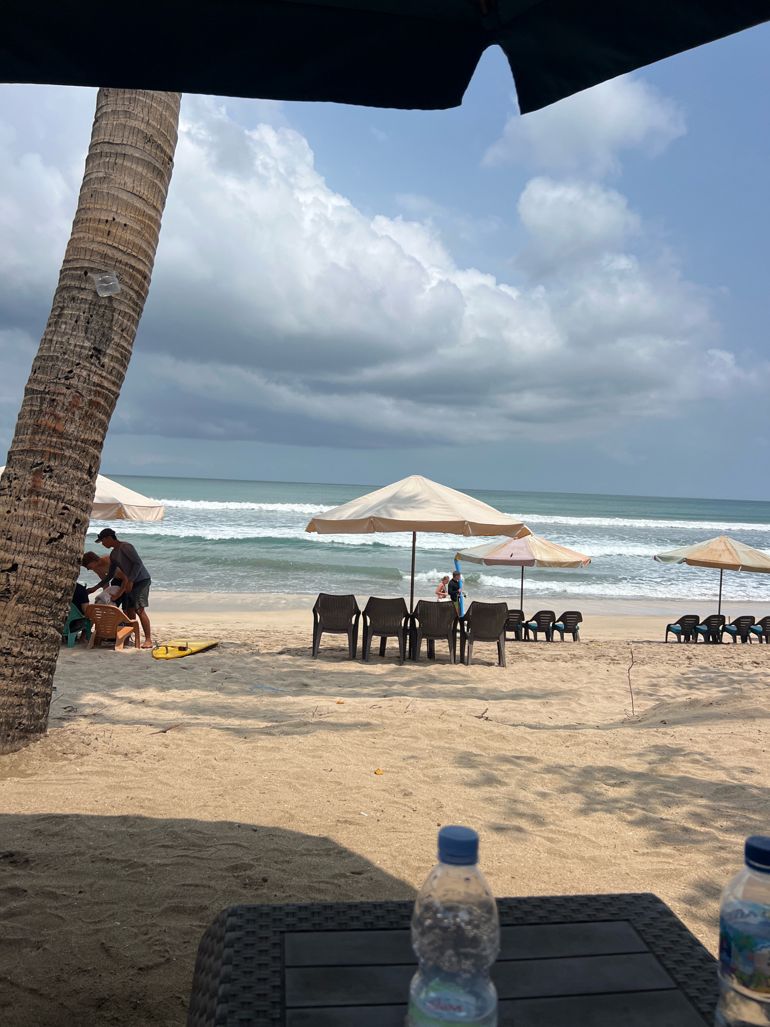 The image depicts a sandy beach in Kuta, Indonesia, on a cloudy day. In the foreground, a dark wicker table holds two plastic water bottles, one with a blue cap and the other with a white cap. A large palm tree trunk is visible on the left side of the frame, with its rough bark textured.
Further into the scene, a group of people are gathered near the water. Two individuals, appearing to be male, are interacting near some beach chairs and an umbrella. One person, wearing a dark long-sleeved shirt and shorts, is leaning over to assist someone else who is partially obscured. A yellow surfboard rests on the sand nearby. Several beach umbrellas are set up along the shoreline, with rows of empty lounge chairs beneath them, suggesting a less crowded day. The ocean in the background shows gentle waves rolling in. The sky is a mix of grey clouds and patches of blue, indicating an overcast but potentially bright day. The sand is a light tan color, with some scattered debris.