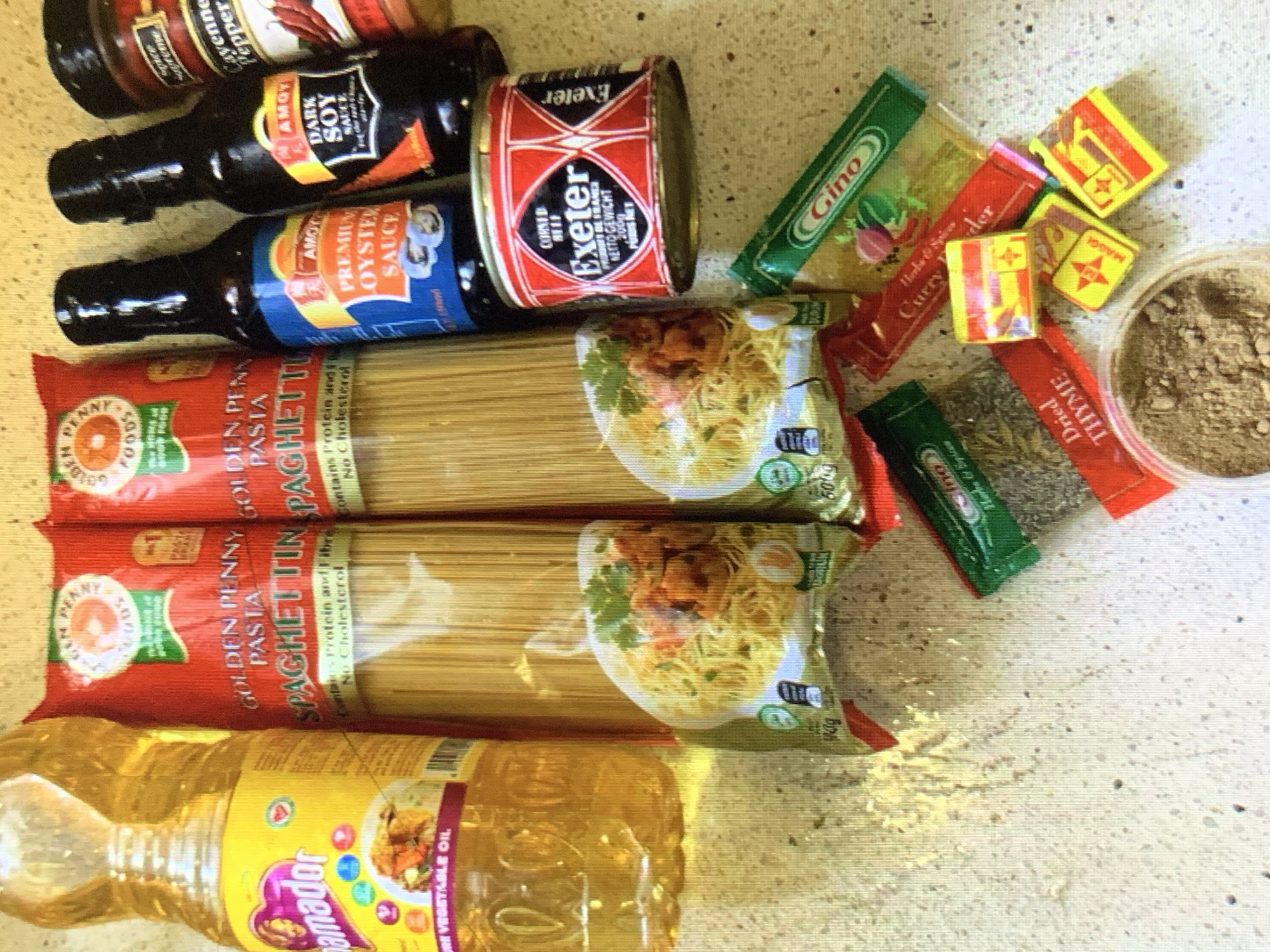 The image shows an assortment of food items on a light-colored surface. These include: two packages of Golden Penny Spaghetti, a bottle of Amador vegetable oil, two bottles of Amoy sauces (Dark Soy Sauce and Premium Oyster Sauce), a can of Exeter corned beef, a container of ground spices, and various seasoning packets (Gino, Curry powder, Dried Thyme, and Maggi cubes). The location of Rafin Par, Nigeria, is not indicated in the image. There are no people depicted in the image.