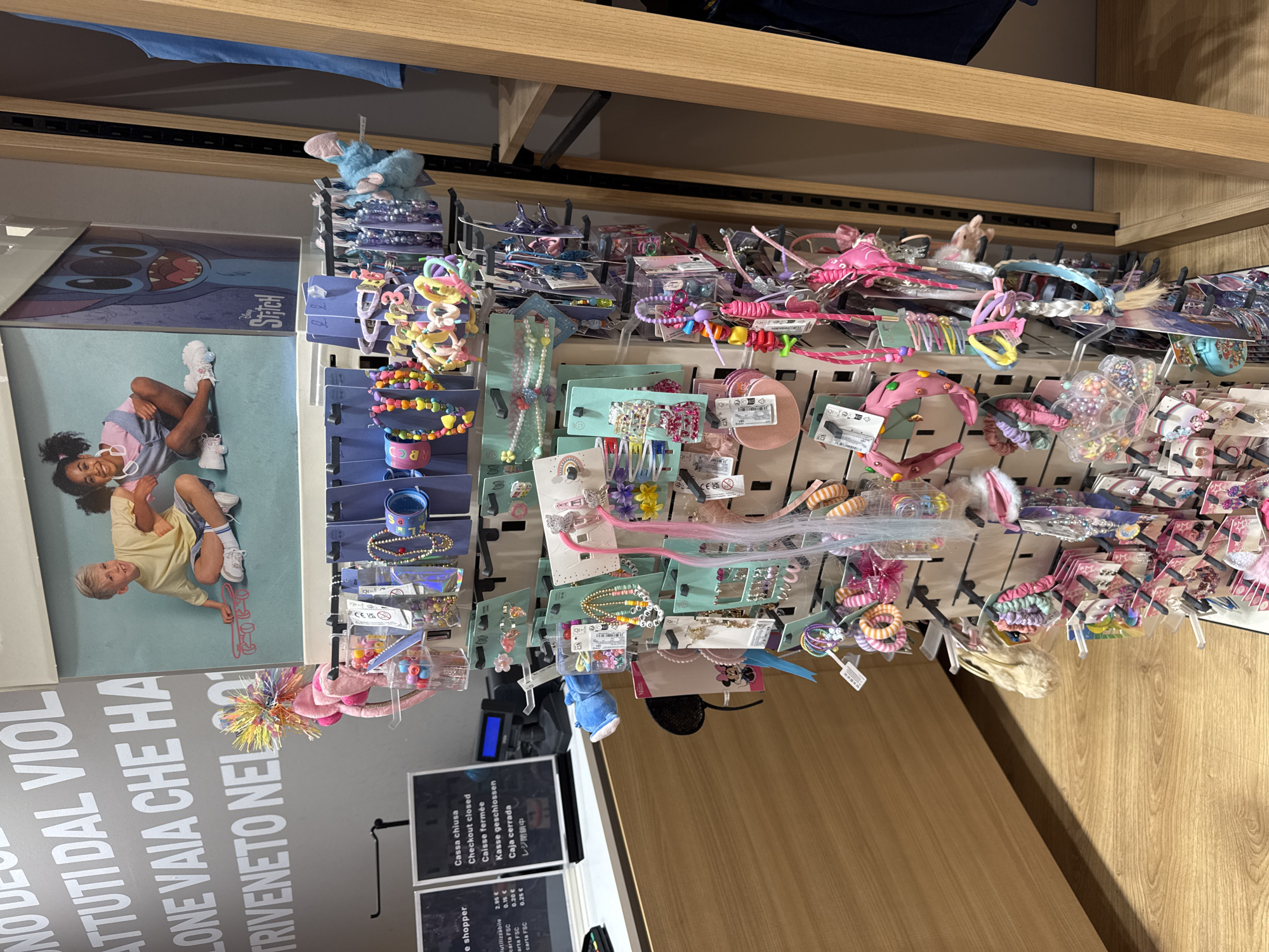 The image shows a retail display of children's hair accessories and jewelry in a store, likely a clothing or accessory shop. The display is a vertical rack with multiple shelves and hooks holding various items such as headbands, hair clips, bracelets, and necklaces. The accessories are colorful and often feature cartoon characters or themes appealing to children, such as Stitch from Disney.
In the background, there is a poster featuring children and the character Stitch. To the left of the display, there is a counter with a cash register and signage indicating "Cassa chiusa" (Closed Checkout), "Checkout closed," "Caisse fermée," and "Kasse geschlossen" in multiple languages, suggesting this area is not currently open for transactions. There are also price lists visible on the counter.
The overall environment appears to be a brightly lit retail space with wooden flooring and a wooden shelving unit on the right side of the frame. The image is taken from a medium distance, capturing a significant portion of the display and its surroundings.
The location is identified as being in Carini, Italy, though this specific detail is not visually ascertainable from the image itself.