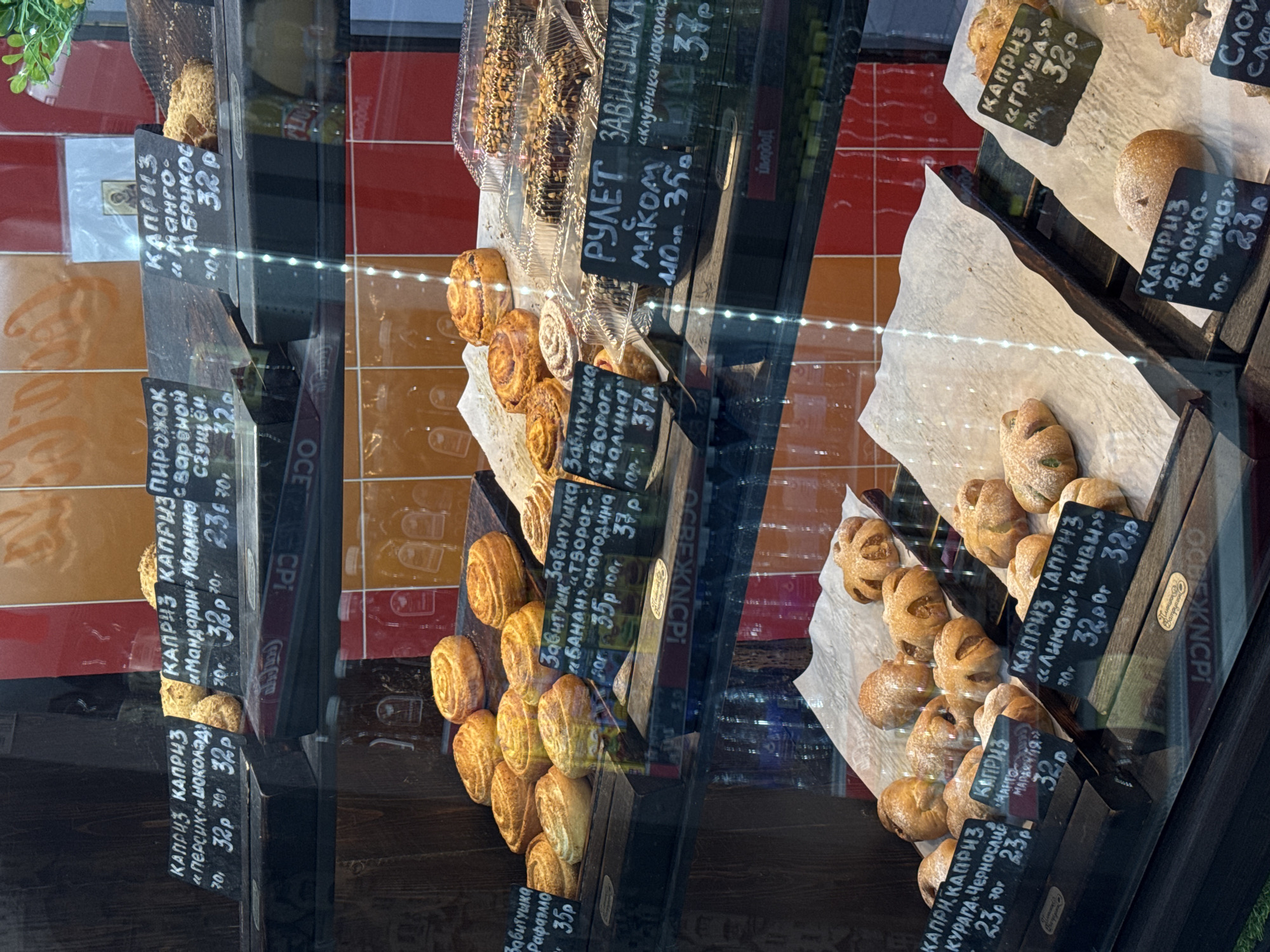 This image captures a vibrant display of baked goods inside a retail setting, likely a bakery or a food section of a store, seen through a reflective glass case. The main subjects are numerous pastries, buns, and rolls, meticulously arranged on wooden trays and shelves.
The scene showcases a variety of baked items, each with a price tag indicating its name and cost in rubles. The dominant type of pastry is labeled "КАПРИЗ" (Caprice), with various fillings such as "Манго-АБРИКОС" (Mango-Apricot) for 32P, "ГРУША" (Pear) for 32P, "ЯБЛОКО-Корица" (Apple-Cinnamon) for 23P, "Мандарин Малина" (Mandarin Raspberry) for 32P, "Персик Шоколад" (Peach Chocolate) for 32P, "ЛИМОН КИВИ" (Lemon Kiwi) for 32P, and "КУРАГА Чернослив" (Dried Apricot Prune) for 23P. Another popular item is "Завитушка" (Curl), available with fillings like "ТВОРОГ" (Cottage cheese) for 37P, "Рафаэлло" (Raffaello) for 35P, "БАНАН СМОРОДИНА" (Banana Blackcurrant) for 35P, and "Клубника шоколад" (Strawberry Chocolate) for 37P. There's also a "РУЛЕТ с МАКОМ" (Poppy seed roll) priced at 35P, and a "ПИРОЖОК с варёной Сгущёнкой" (Pirozhok with boiled condensed milk) for 32P.
The activity taking place is the presentation of these pastries for sale. The baked goods are positioned to entice customers, with some dusted with powdered sugar or topped with nuts. The background behind the display consists of red and orange tiles, adding a contrasting color to the scene. The reflections on the glass indicate an indoor environment, possibly reflecting other parts of the shop or a plant. The text on the price tags is in Russian, suggesting the location is in a Russian-speaking region. No specific time of day or weather can be discerned.