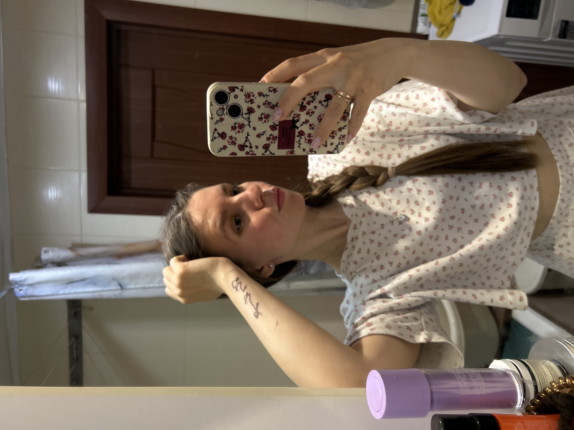 A young woman is captured in a mirror selfie, holding a smartphone with a cream-colored floral case featuring red and pink roses. Her fingernails are painted pink with white polka dots, and she wears a gold-colored ring. She has long, dark brown hair styled in a braid over her left shoulder and is wearing a white short-sleeved top adorned with small pink and red floral patterns. On her left forearm, a tattoo in cursive script is visible, which appears to read "Good energy."
The reflection in the mirror reveals an indoor setting, likely a bathroom or dressing area. A dark wooden door frame is prominent in the background, along with a white tiled wall. To the left, a white and blue object, possibly a utility shelf or a clothes drying rack, is reflected. The bottom right corner of the mirror shows various items on a surface, including a purple-capped bottle and other containers. The lighting suggests daytime. The woman gazes directly into the camera lens with a neutral expression. The location context provided is Klin, Russia.