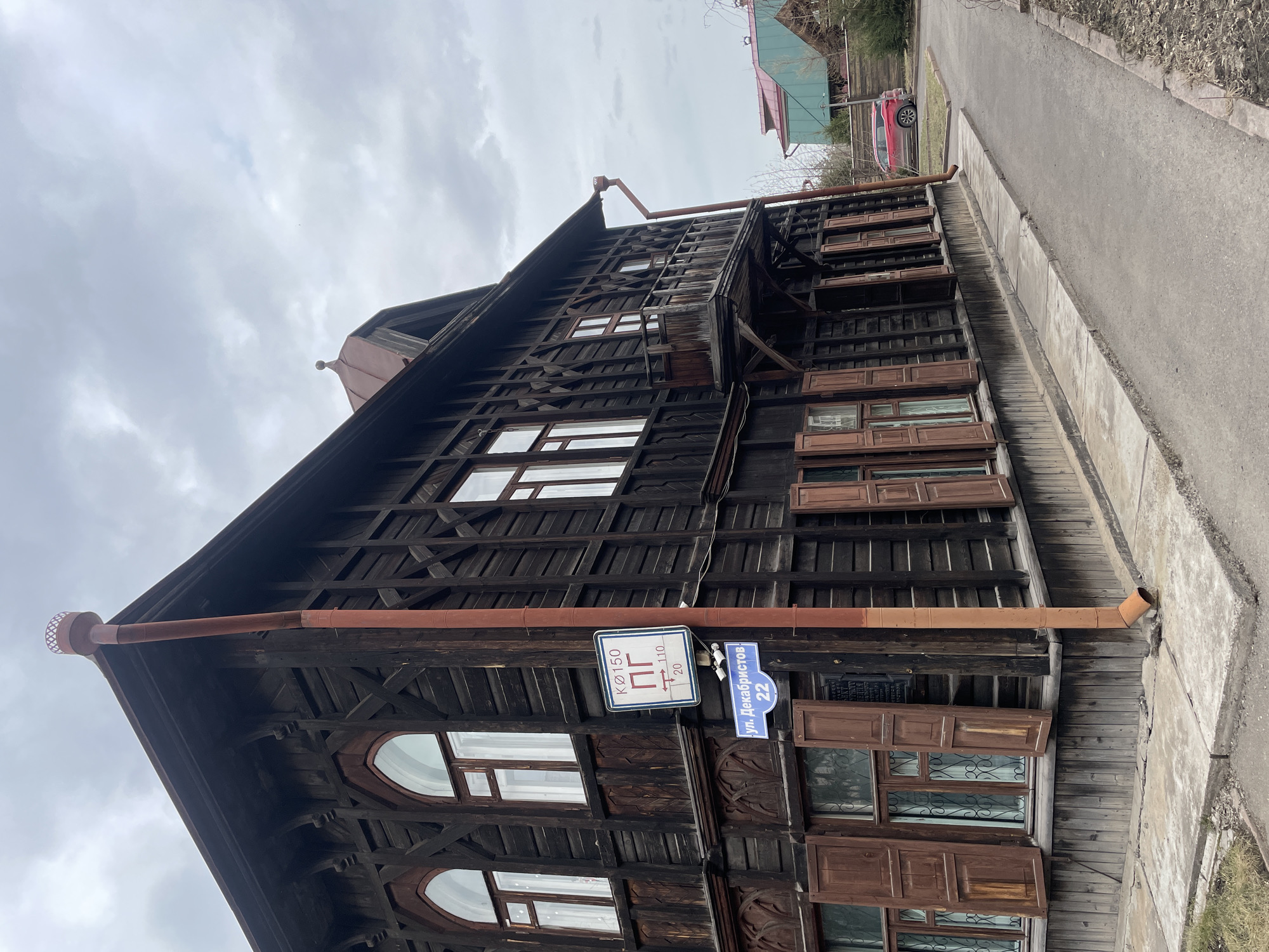 A large, dark wooden building, likely a historic residence, is the central subject of the image, seen under an overcast sky in Krasnoyarsk, Russia. The building features ornate wooden patterns on its facade and multiple windows, some with closed or partially open brown wooden shutters. A rusty brown gutter system is visible along the roofline and descending the building's side.
Mounted on the building's facade, two signs are prominent. A blue street sign clearly states "ул. Декабристов" (Decembrists Street) and the house number "22". Below it, a white sign with a blue border displays "КØ150", the Cyrillic letters "ПГ" (likely indicating a fire hydrant), and directional arrows with numerical indicators "↑110" and "→20", which typically denote distance and direction to the hydrant.
To the right of the building, a paved sidewalk runs alongside a grey asphalt road. In the distance along the road, a red car is partially visible, parked near another building with a green roof. The ground beside the sidewalk appears to have dry grass or vegetation. The sky is completely cloudy, suggesting an overcast day. No people or specific activities are depicted in the static street scene.