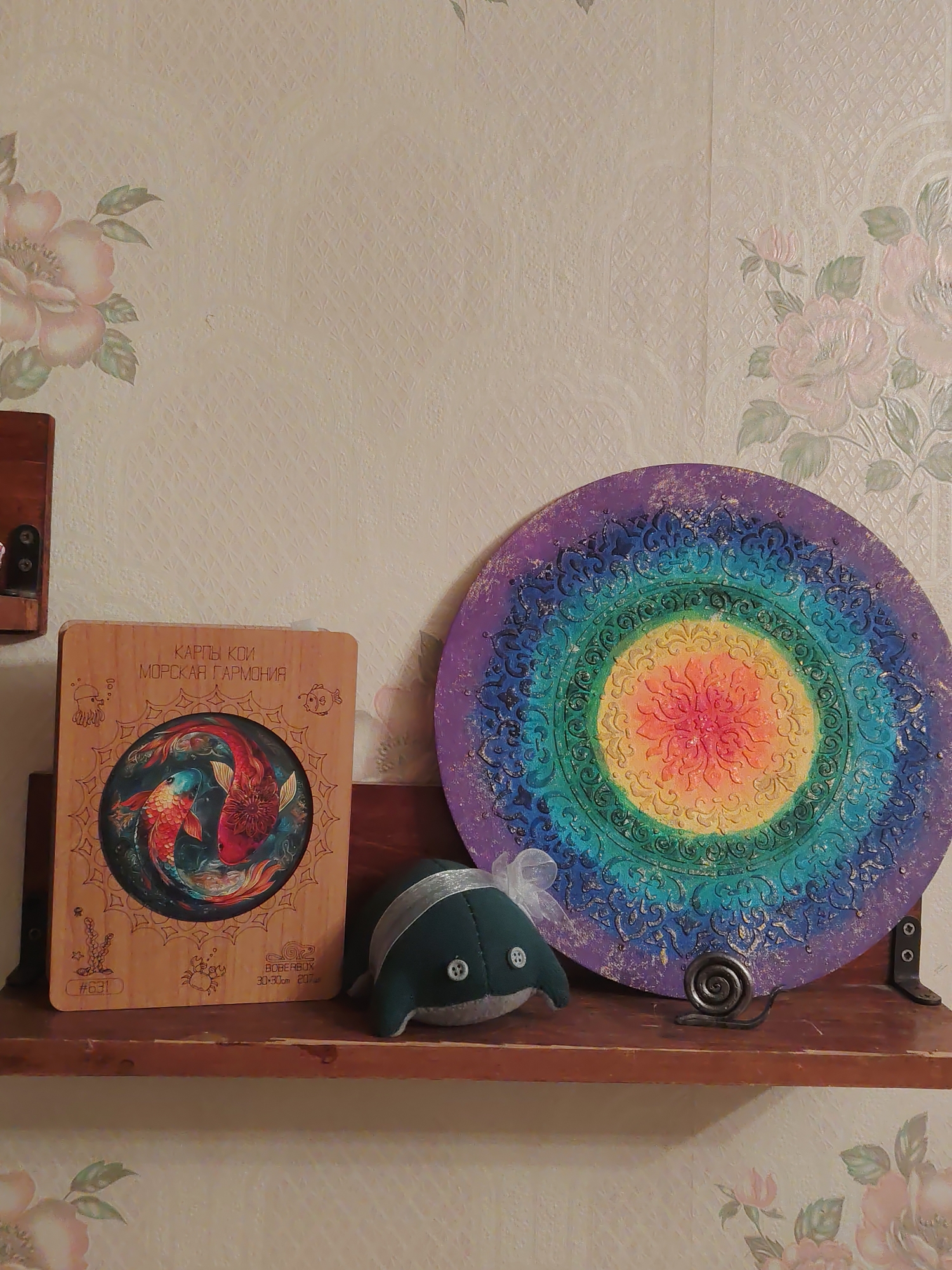 The image shows a shelf against a floral wallpaper. On the shelf, from left to right, is a wooden plaque with an illustration of koi fish and Russian text, a small plush toy shaped like a whale with a white ribbon tied around its head, and a circular mandala-like art piece with rainbow colors and intricate patterns. To the right of the whale toy, on the shelf, is a small metal snail sculpture. The shelf appears to be made of dark wood. The overall scene suggests a domestic setting. There is no discernible information about the city of Saratov, Russia, within the image.