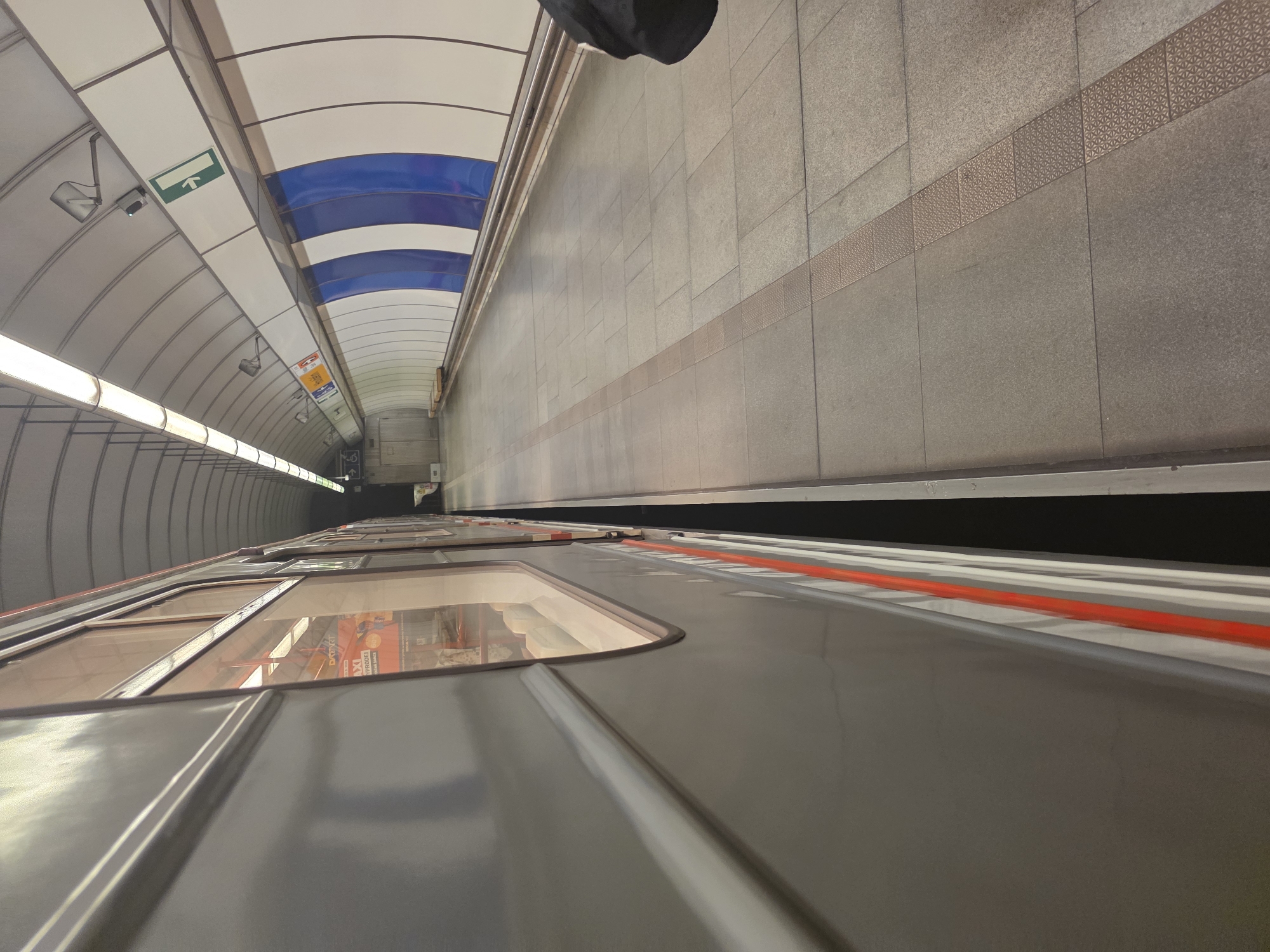 The image shows a subway platform and train car in an underground station. The station has a curved ceiling with blue and white stripes. The platform is tiled in gray and beige. A train car is visible in the foreground, with its windows reflecting the platform. The station appears to be clean and well-maintained. The location is possibly London, United Kingdom.