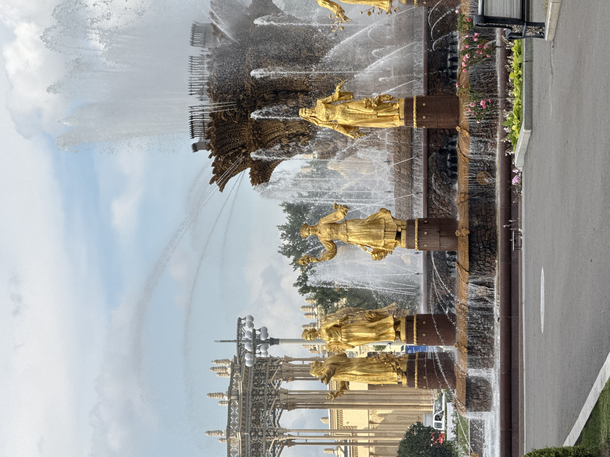 The image shows a view of the "Friendship of Peoples" fountain at VDNKh (Exhibition of Achievements of National Economy) in Moscow, Russia. The central focus is the large, ornate fountain, characterized by its golden statues representing different Soviet republics. Water streams cascade from the fountain's upper tiers, creating a dynamic and majestic display. In the background, architectural elements of VDNKh, including pavilions and decorative structures, are visible. The sky is partly cloudy, with a mix of blue sky and white clouds. The foreground features a paved area, landscaping with greenery and pink flowers, and a park bench. Several golden statues of women in flowing dresses, each holding symbolic items like flowers or wheat, stand prominently around the fountain's base. The overall scene depicts a sunny day at a popular Moscow landmark, highlighting the grandeur of Soviet-era architecture and art.