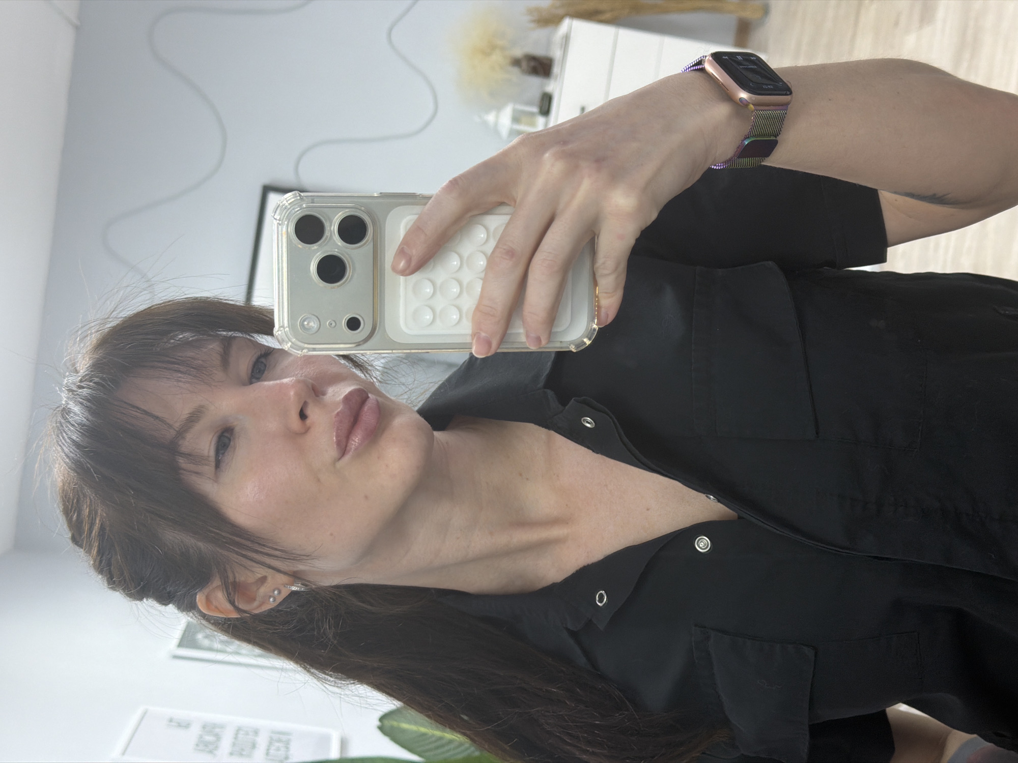 A woman in a black collared shirt is holding up a cell phone in her right hand, taking a selfie. She is wearing an Apple Watch with a colorful metal band. The phone is an iPhone with three camera lenses and a clear case with a white grip on the back. The background shows a blurry room with a white dresser, a framed picture on the wall, and possibly some plants. There is no direct indication that this image was taken in Perm, Russia.
