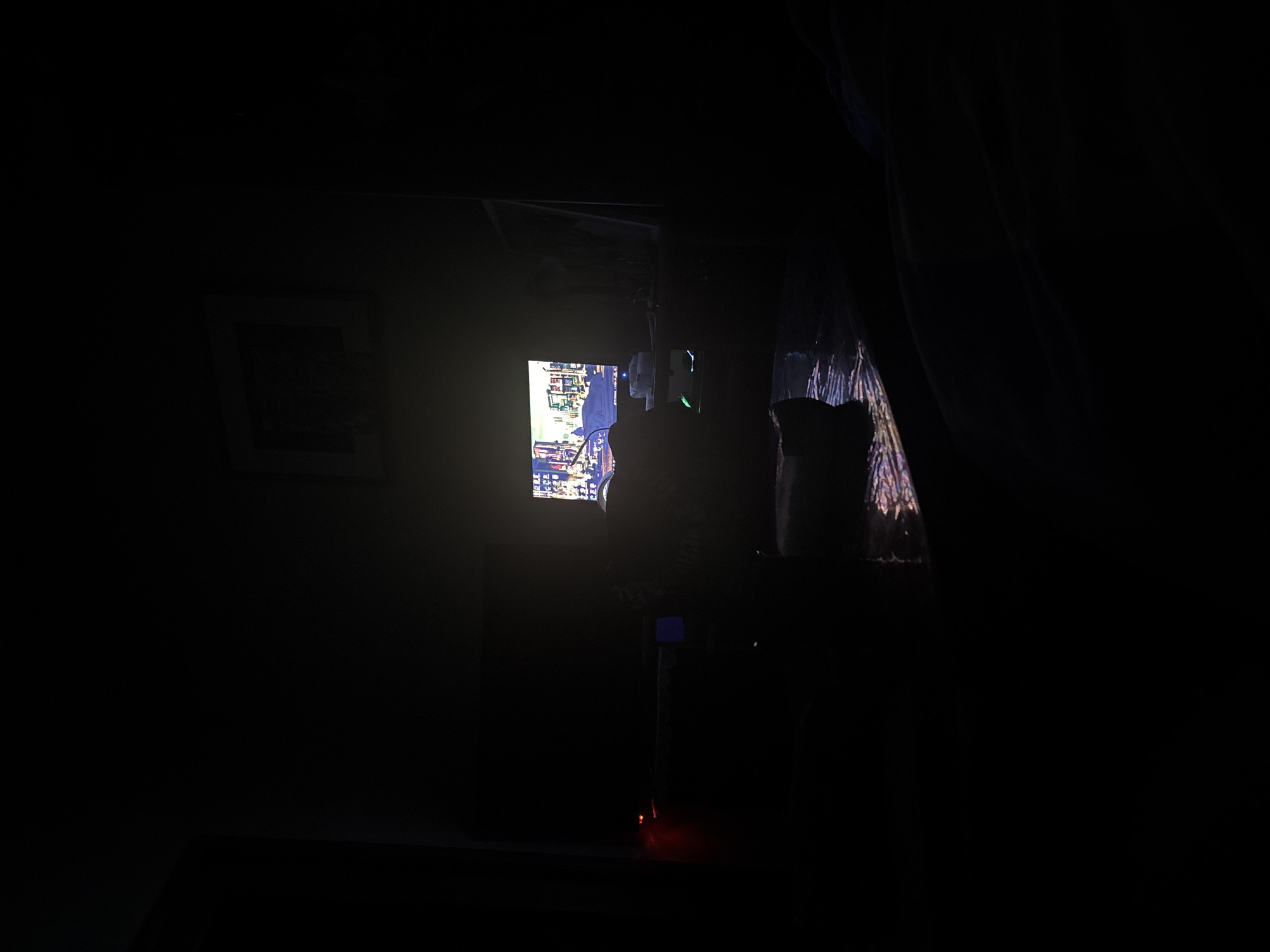 A low-lit room with a framed picture on the left wall. A television displays an image of Yerevan, Armenia. A curtain hangs to the right of the television. On a table or shelf in front of the television, there's what appears to be a container or object, its shape obscured by darkness. A light emitting from the bottom of a stand is at the bottom of the frame.
