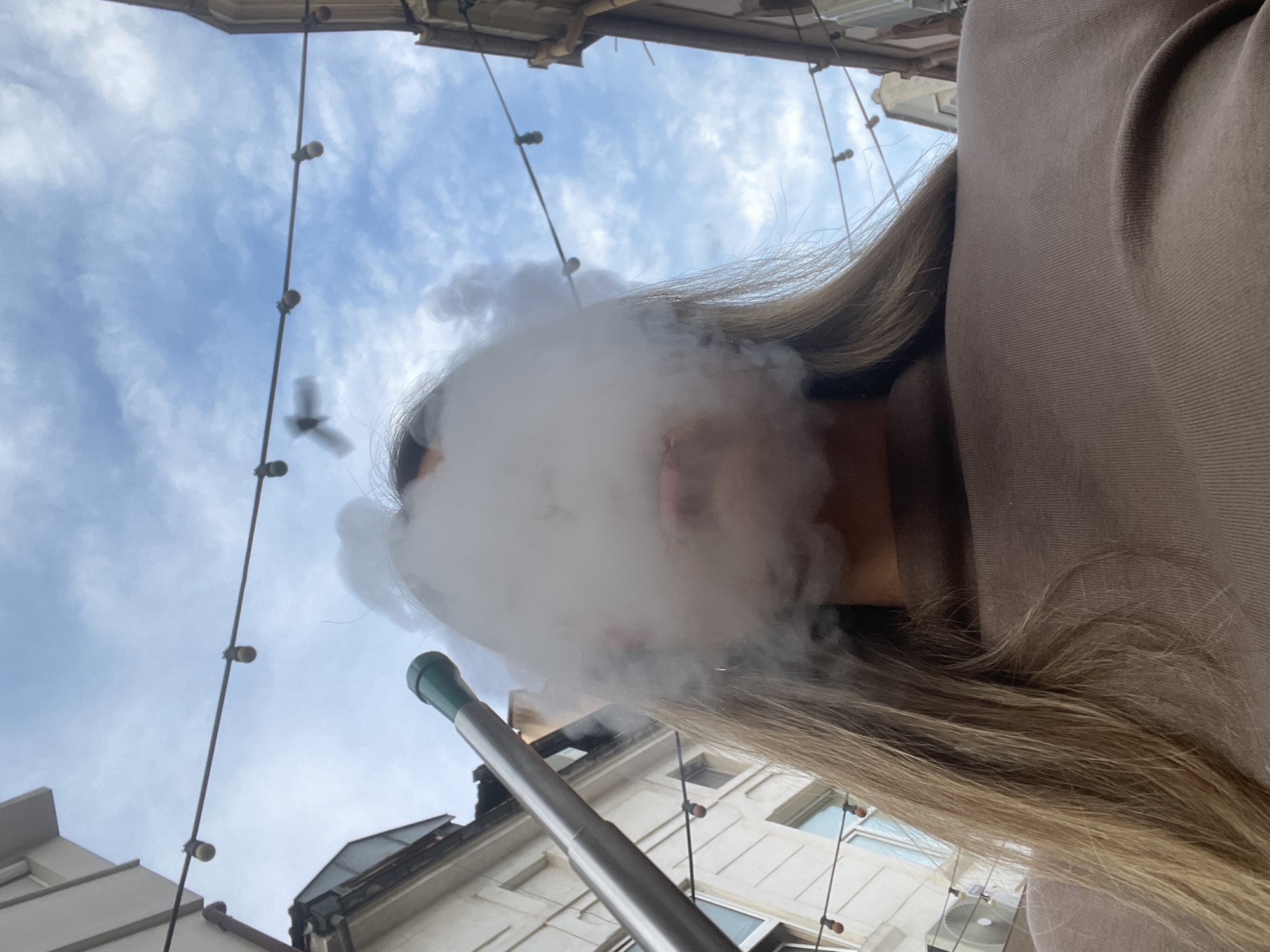 A low-angle, medium shot captures a young adult in the foreground, exhaling a plume of white smoke, obscuring much of their face. The smoke is illuminated by natural light, creating a hazy effect around the subject's mouth and nose. A portion of their long, blonde hair is visible to the left of the frame, framing the lower part of their face. They are wearing a brown, long-sleeved shirt. In the background, a metallic hose with a green tip is partially visible on the left side, suggesting the use of a hookah. Above, a string of lights with small bulbs hangs across the frame, with buildings on either side and a cloudy blue sky in the distance. The overall scene appears to be outdoors in an urban setting.