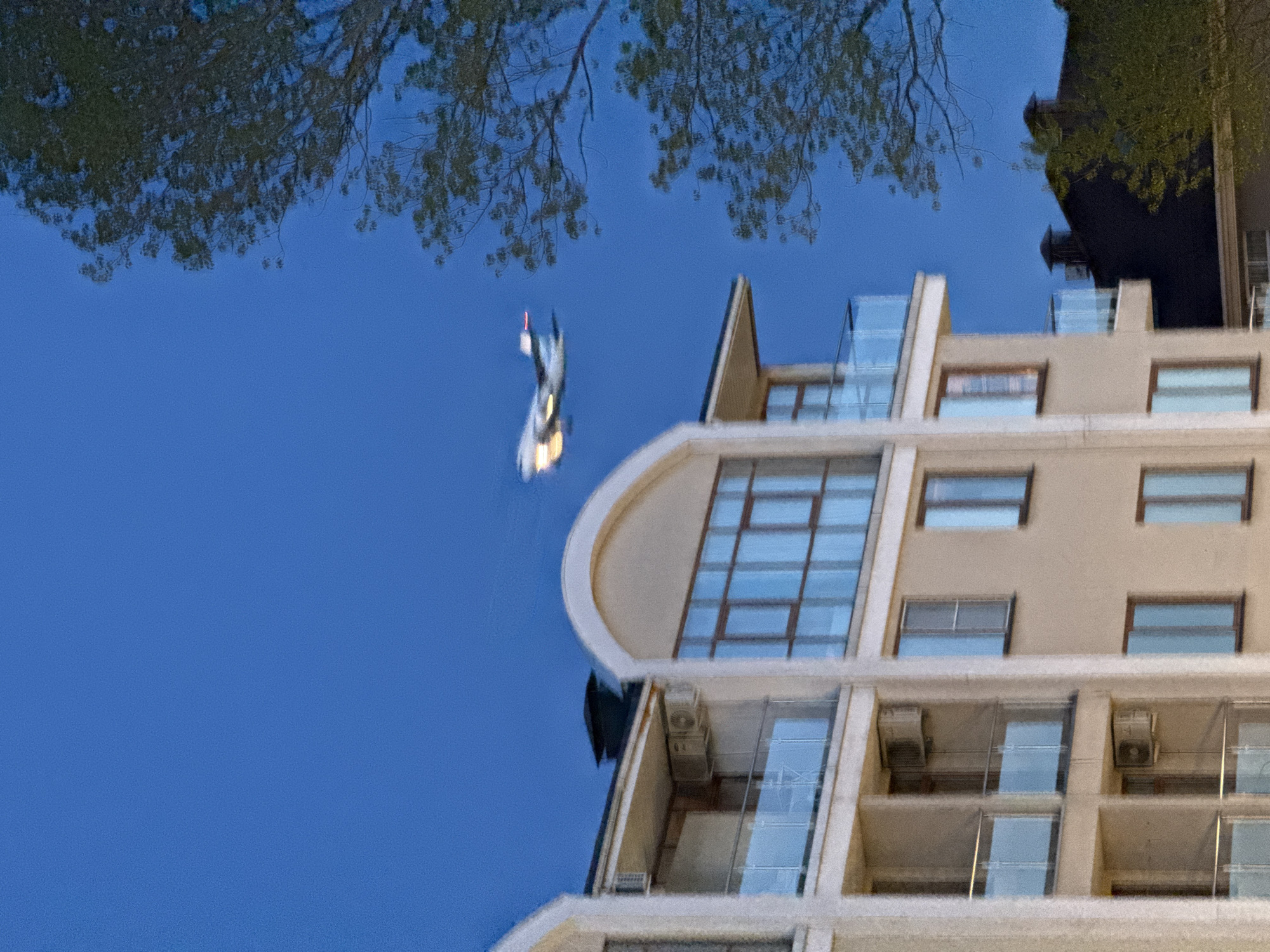 A modern, multi-story building, likely an apartment complex or hotel, stands prominently in the image. It features a light, possibly beige or cream-colored facade, numerous windows with brown frames, and glass-railed balconies equipped with visible air conditioning units. A distinctive architectural detail includes a curved section with large windows on an upper floor.
The image captures a serene outdoor scene, dominated by a clear, deep blue sky and the reflection of surrounding elements. The upper part of the frame is filled with the vivid reflection of tree branches and leaves silhouetted against the blue. This highly reflective surface, likely a calm body of water, also captures the blue of the sky with remarkable clarity.
A striking and unusual detail is an elongated, bright, and blurry object reflected against the blue. This object appears to be a human figure, dressed in white or reflective clothing, with a distinct red tip or element. Its posture and position suggest it is in motion, possibly diving into the water or falling, creating a dynamic point of interest within the otherwise tranquil scene.
The time of day appears to be daytime, perhaps early morning or late afternoon, indicated by the rich blue of the sky and the soft lighting. The weather is clear, with no clouds visible. The presence of a modern building and implied body of water aligns with the location context of Sochi, Russia, likely along its coast. There is no visible text in the image.