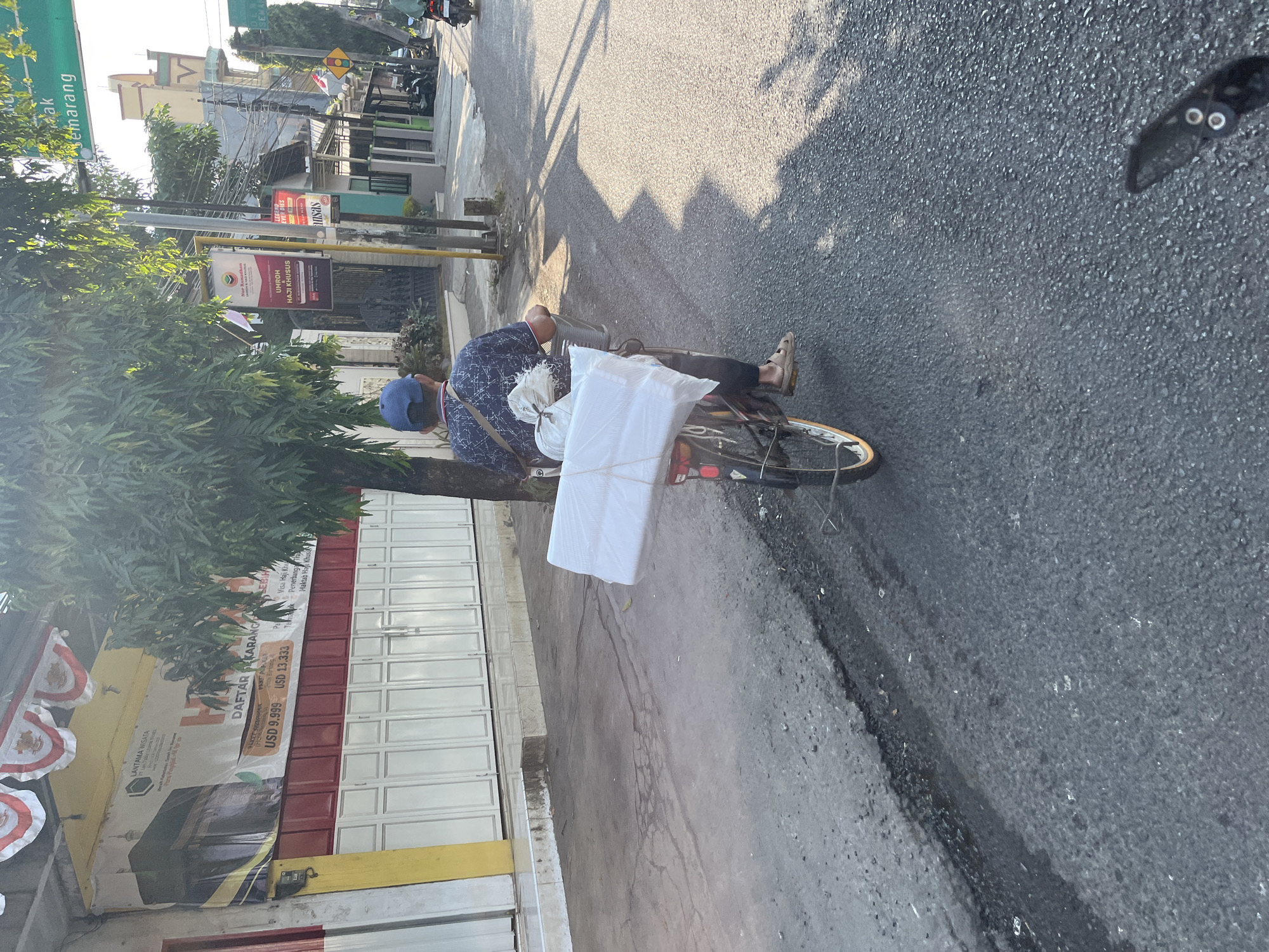 In Kalinyamatan, Indonesia, a man wearing a blue baseball cap and a patterned blue shirt is riding a bicycle on a paved road. He is carrying a large, white, wrapped object secured to the back of his bicycle. The object appears to be rectangular and is tied down with rope. The man is wearing sandals and has a bag slung over his shoulder. In the background, there are buildings with red and white doors, a tree, and other parked vehicles. The sun is shining, casting long shadows on the road. A green street sign with Indonesian text is visible in the upper right portion of the image.
