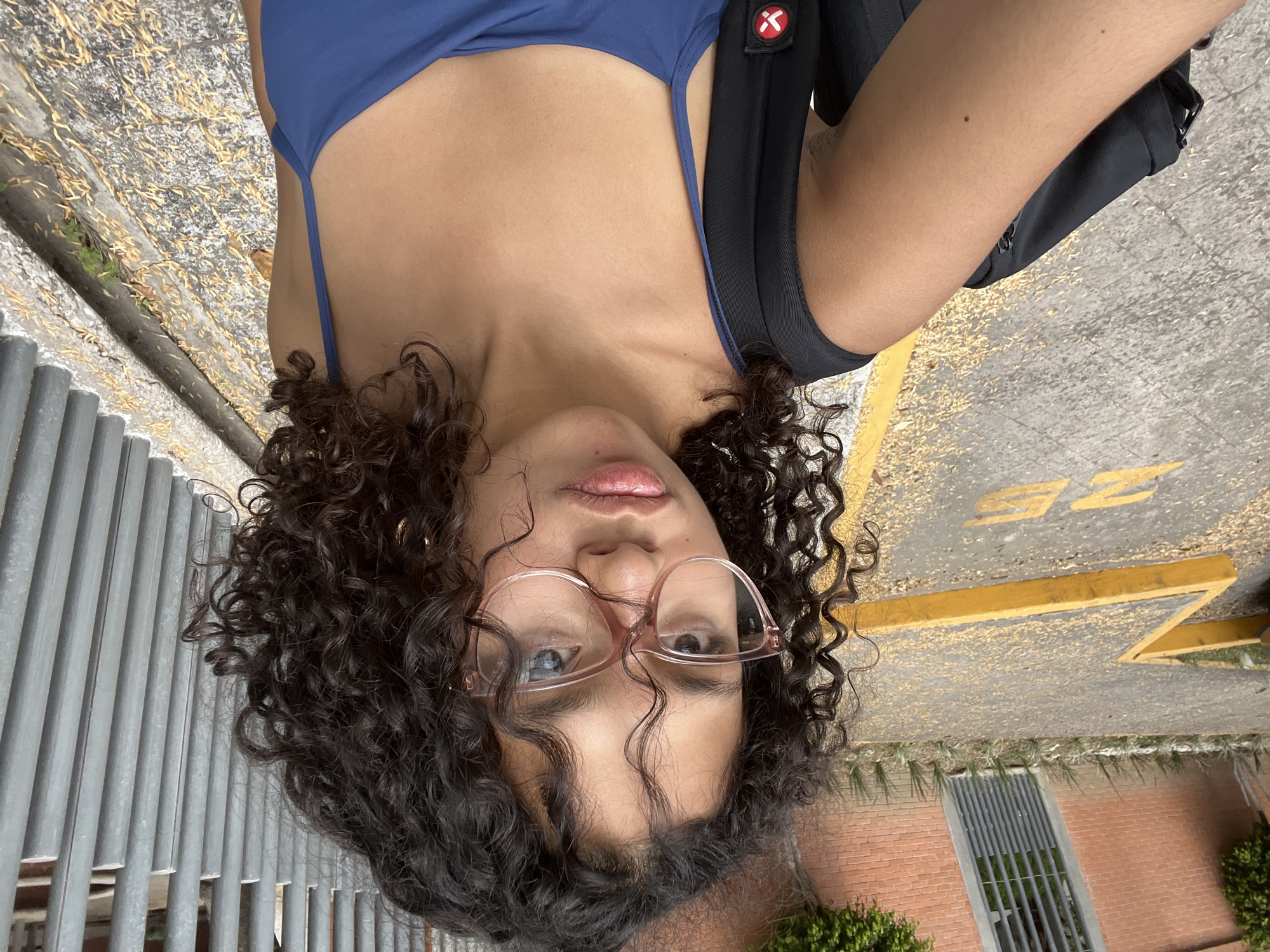 A person with curly dark hair and pink glasses is shown from a low angle. They are wearing a blue tank top with a black backpack. The background is a concrete surface with yellow markings, and a metal fence runs along the left side. In the lower right, there is a brick wall with greenery.