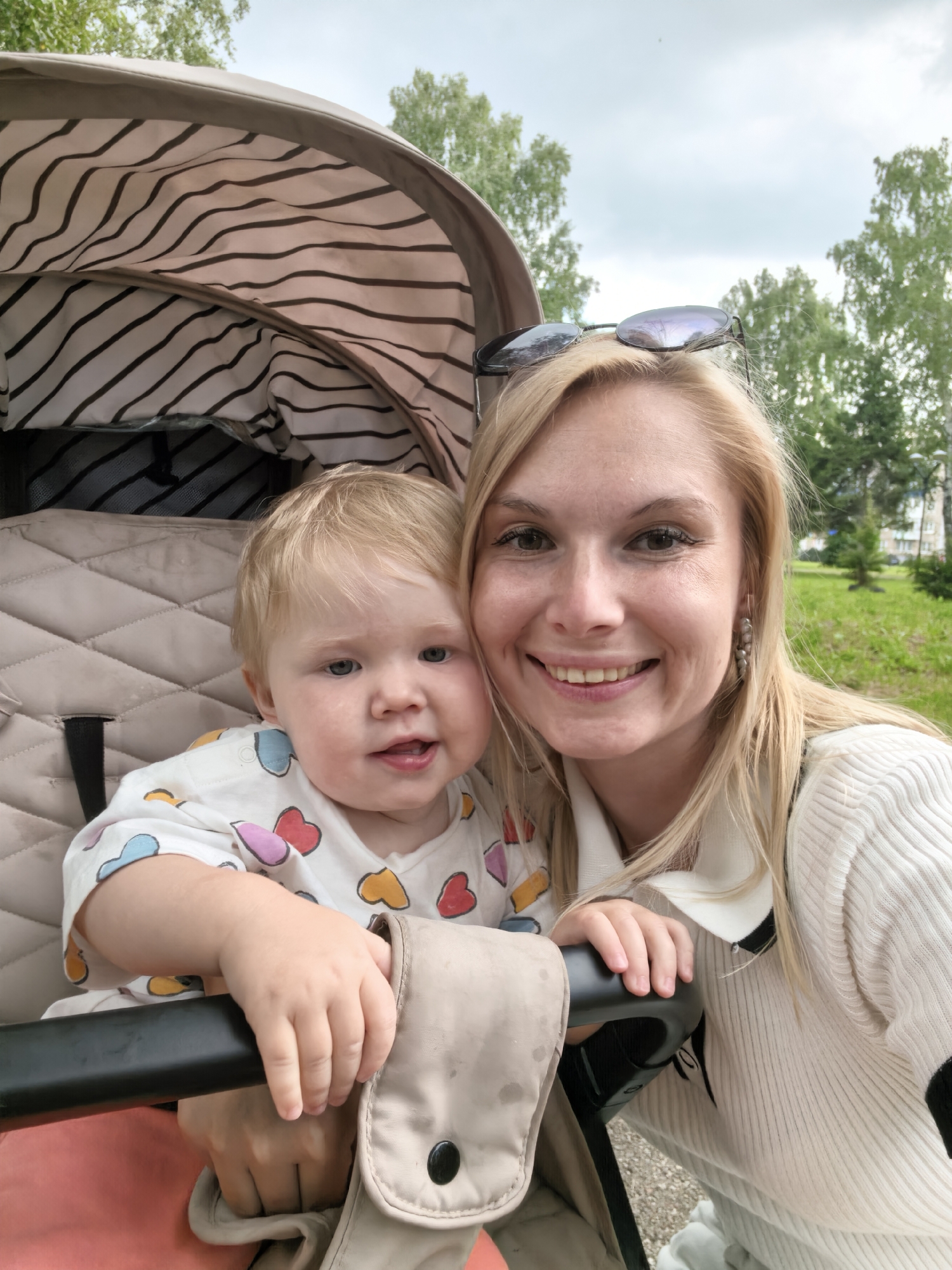 In a city park in Sterlitamak, Russia, a blonde woman and a young child are captured in a close-up selfie. The woman, wearing a white ribbed sweater and sunglasses perched on her head, smiles warmly at the camera. Beside her, a baby with blonde hair sits in a stroller, dressed in a white onesie adorned with colorful heart prints. The stroller's canopy, lined with black and white stripes, is visible in the background. The setting suggests a pleasant day outdoors, with trees and greenery forming a soft backdrop under a cloudy sky.