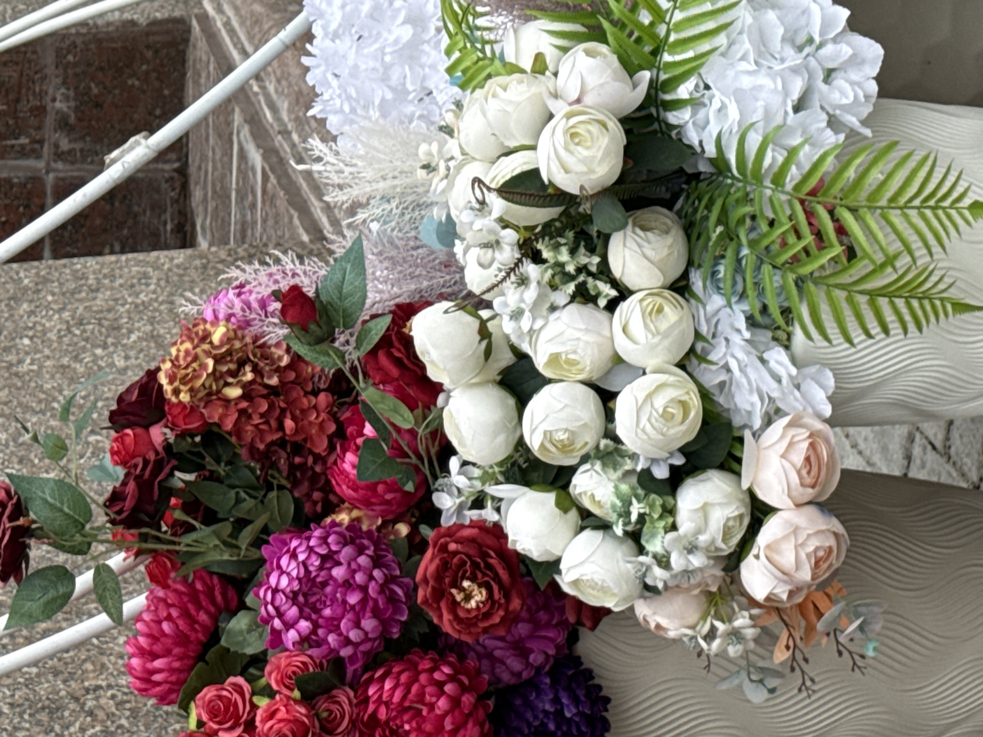 This is a medium shot, eye-level, outdoor, close-up photograph of a flower arrangement featuring a variety of artificial flowers. The arrangement is composed of large, ruffled, pink and magenta peonies or chrysanthemums, deep red roses, smaller burgundy and red flowers resembling hydrangeas, and clusters of white, rose-bud-shaped flowers. There are also green leaves and some wispy, light-colored foliage interspersed throughout. The flowers are arranged in two white, textured vases, one of which is partially visible on the left side of the frame, and the other is to the right, out of focus. The background consists of a textured, possibly stone or brick, wall and a glimpse of a metal railing. The overall impression is of a decorative floral display, likely for an event or an establishment. There are no people or animals present in the image. The image does not provide any specific visual cues to indicate a location within Moscow, Russia.