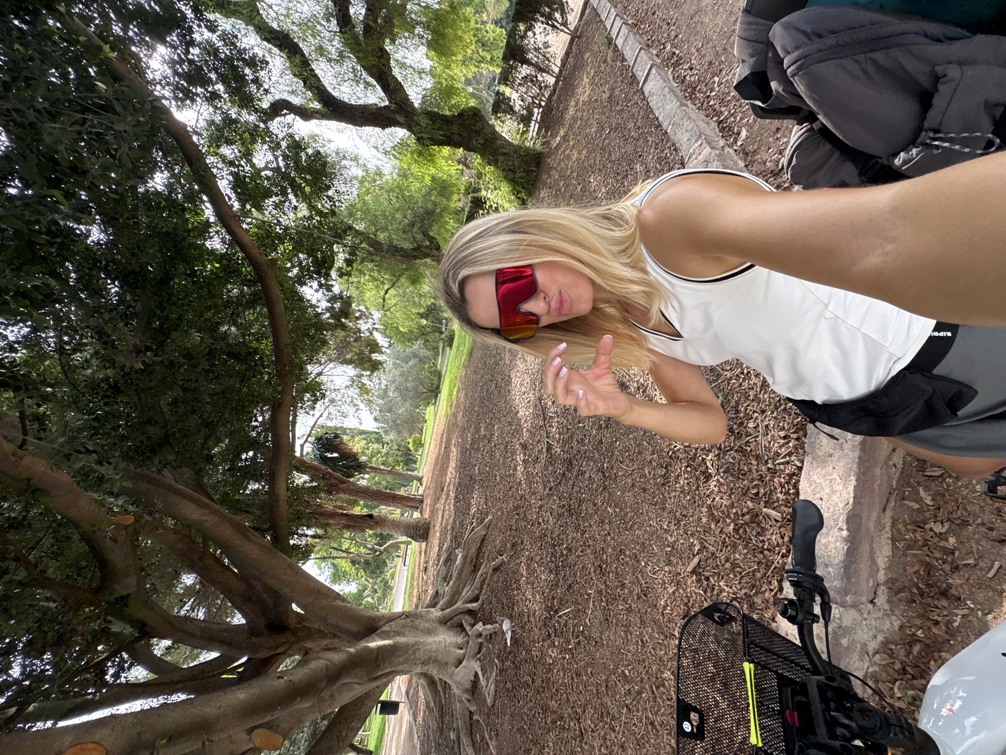 A blonde woman with red-tinted sunglasses is taking a selfie outdoors. She is wearing a white tank top with black trim and grey shorts. She is making a heart shape with her fingers. In front of her is a bicycle with a black basket. The background shows a park with large trees and fallen leaves on the ground. The scene appears to be in Valencia, Spain.