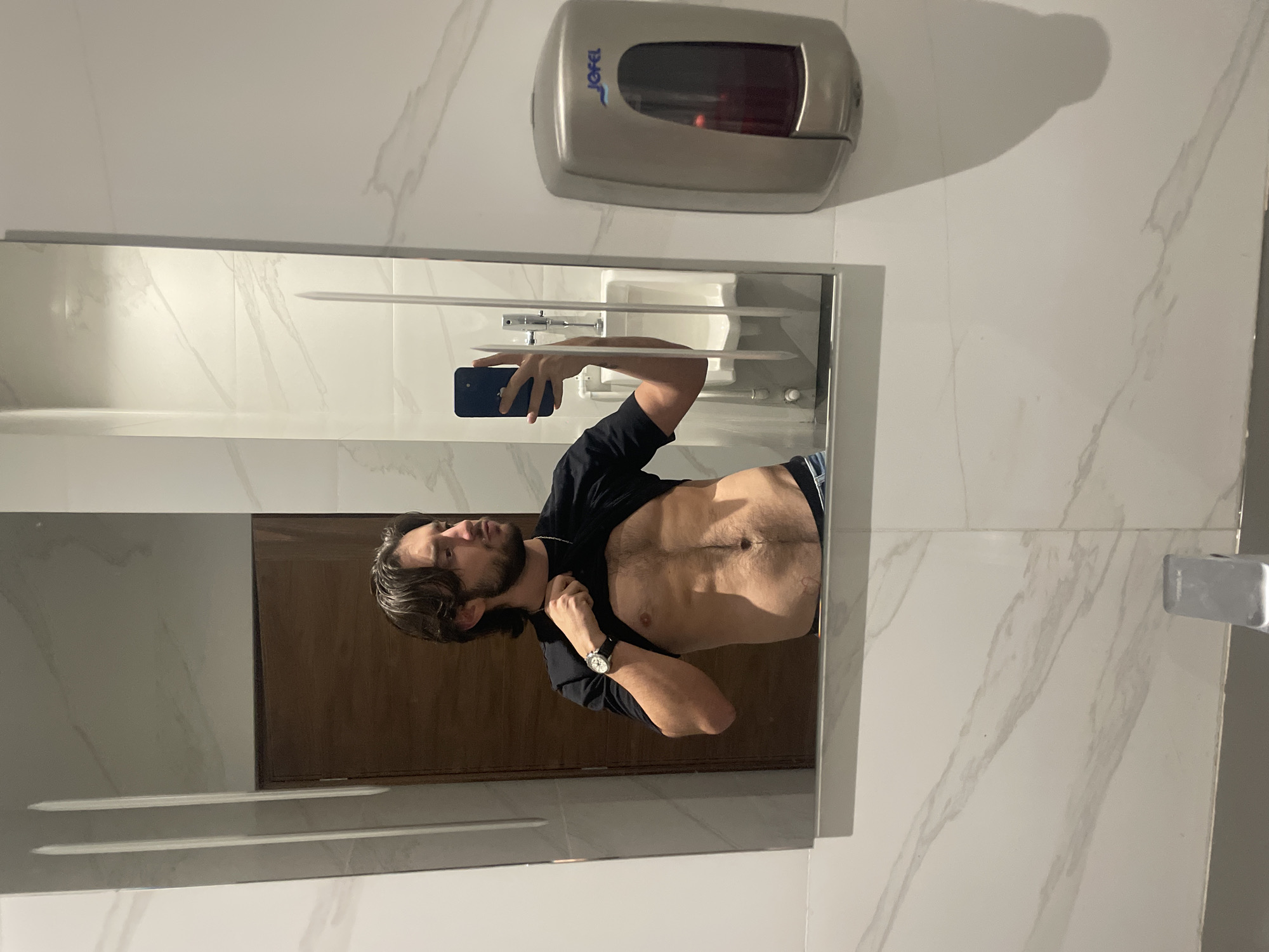 A man is reflected in a mirror in a bathroom. He holds a smartphone in his left hand, presumably taking a photograph. The man is fair-skinned with dark hair and a beard. He is wearing a black shirt, partially unbuttoned, revealing his torso. A watch is visible on his left wrist. Behind him in the mirror are bathroom fixtures, including a sink and a door, possibly made of wood. The wall is covered with white marble tiles. Above the mirror is a "Jofel" branded soap dispenser.
The image does not contain sufficient details to confirm its location in Mexico City.