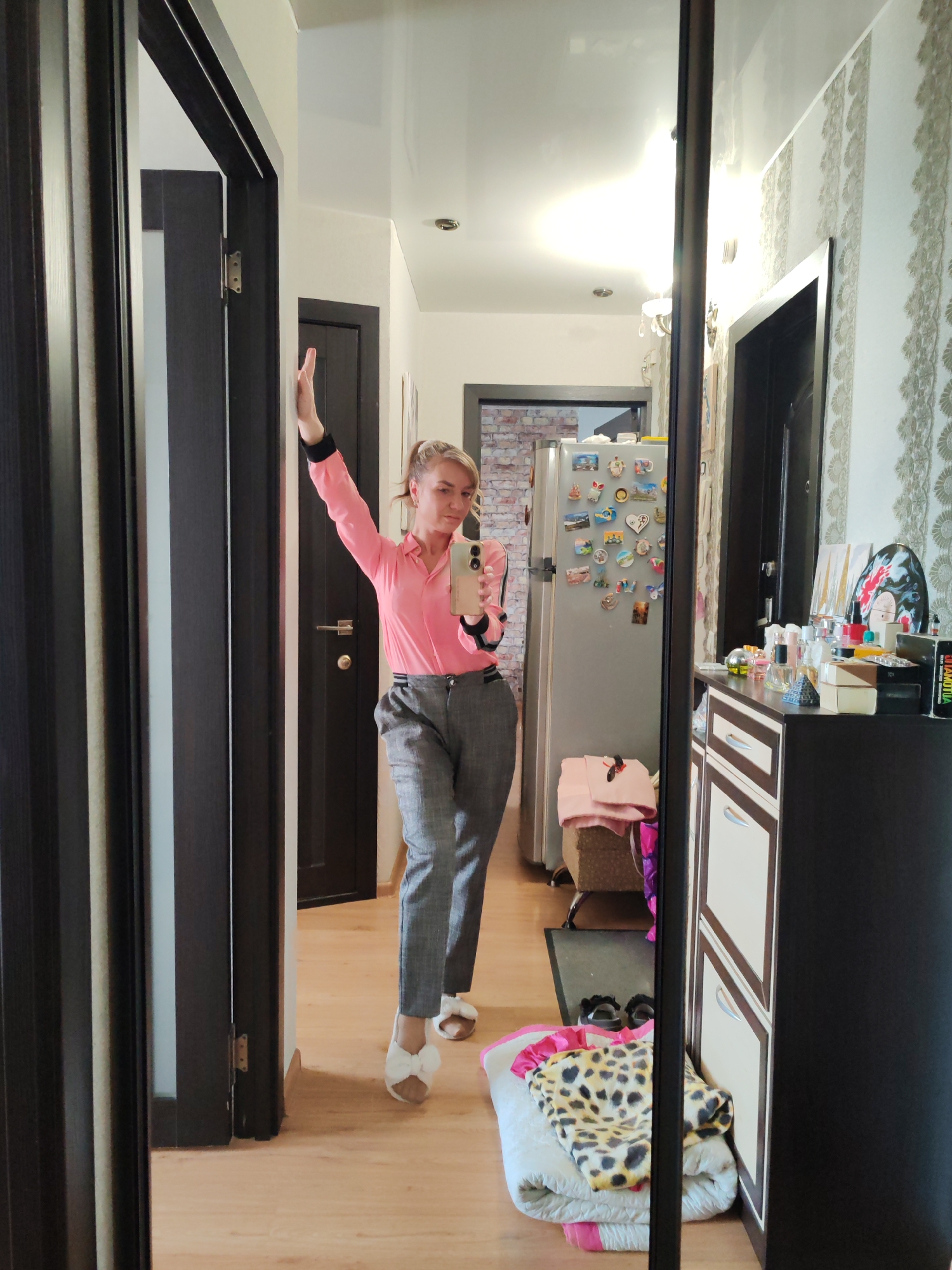 The image shows a full-body selfie of a woman in a pink collared shirt and gray trousers, taken in a hallway. She is standing in front of a dark brown door, with her right arm raised, holding a smartphone. Her left arm is bent at the elbow, with her hand resting on her hip. The hallway has light wood flooring and a white ceiling with recessed lighting. To the left of the woman is a dark brown door frame and another dark brown door. To the right, a large mirror reflects the scene, showing a silver refrigerator covered in magnets, a chest of drawers, and various decorative items. The background suggests an interior residential setting. The image is framed by the dark frame of the mirror. The location is identified as Petropavlovsk-Kamchatsky, Russia.