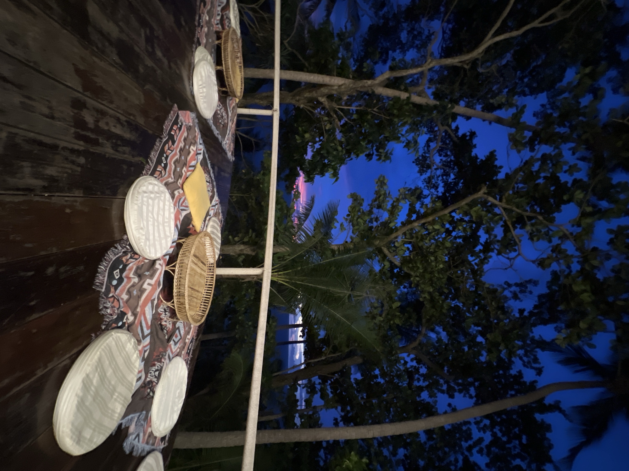 The image depicts an outdoor dining setup on a wooden deck at dusk, likely in Karon, Thailand. A table is set with patterned placemats, white circular platters, and woven baskets. The backdrop is a lush natural scene with large trees and a dark blue twilight sky, suggesting the end of the day. The lighting is dim, hinting at the transition from sunset to night. The overall atmosphere appears serene and relaxed, potentially evoking a sense of vacation or a peaceful evening outdoors. There is no visible text in the image.