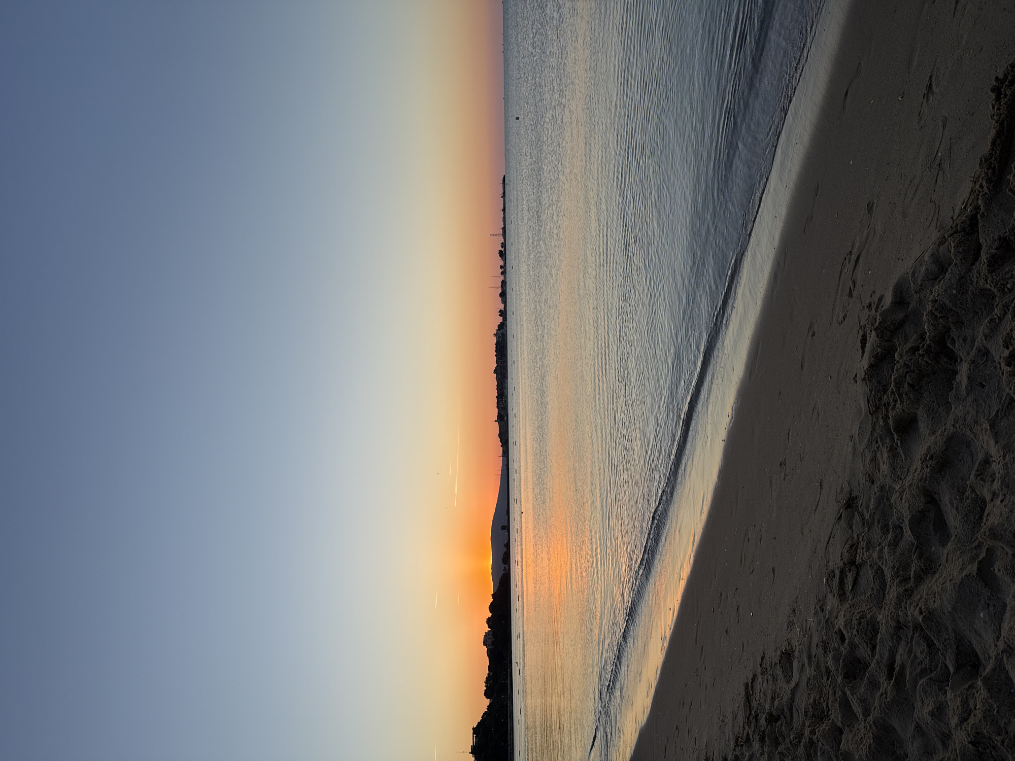 The image shows a serene beach scene in Nesebar, Bulgaria, at sunrise. The sky transitions from a clear, pale blue at the top to a warm orange and yellow gradient near the horizon, indicating the early morning light. The sun is just peeking over a distant hill, casting a vibrant reflection on the calm sea. The water is a deep blue with gentle ripples, lapping at the sandy shore. The foreground features dark, wet sand with footprints and some scattered debris. In the middle ground, the sea is dotted with buoys. The background shows a silhouette of a town on a hill with buildings and structures, likely the historic part of Nesebar. The overall impression is one of tranquility and natural beauty.