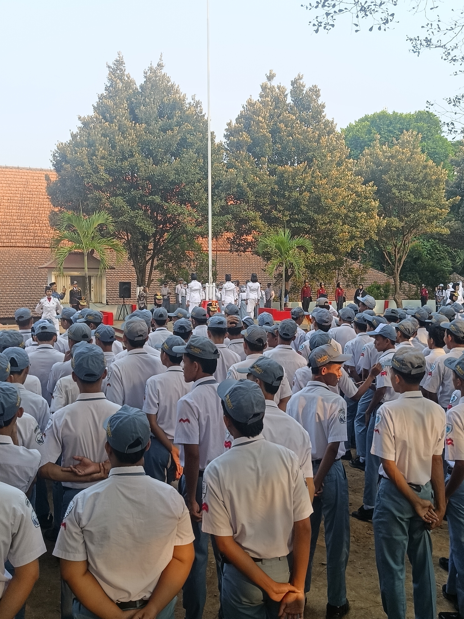The image depicts a gathering of Indonesian students, likely in uniform, standing outdoors in what appears to be a schoolyard or public space in Pengkol, Indonesia. In the foreground, a large group of students in white shirts and gray hats are facing away from the camera, their hands clasped behind their backs. They are standing on a dirt ground. Behind them, a smaller group of individuals in white uniforms, possibly teachers or officials, are positioned near a raised red platform and a flagpole. Trees and a building with an orange tile roof are visible in the background, suggesting a tropical or subtropical environment. The overall scene suggests an organized event, such as a flag ceremony or assembly, taking place during daylight hours.