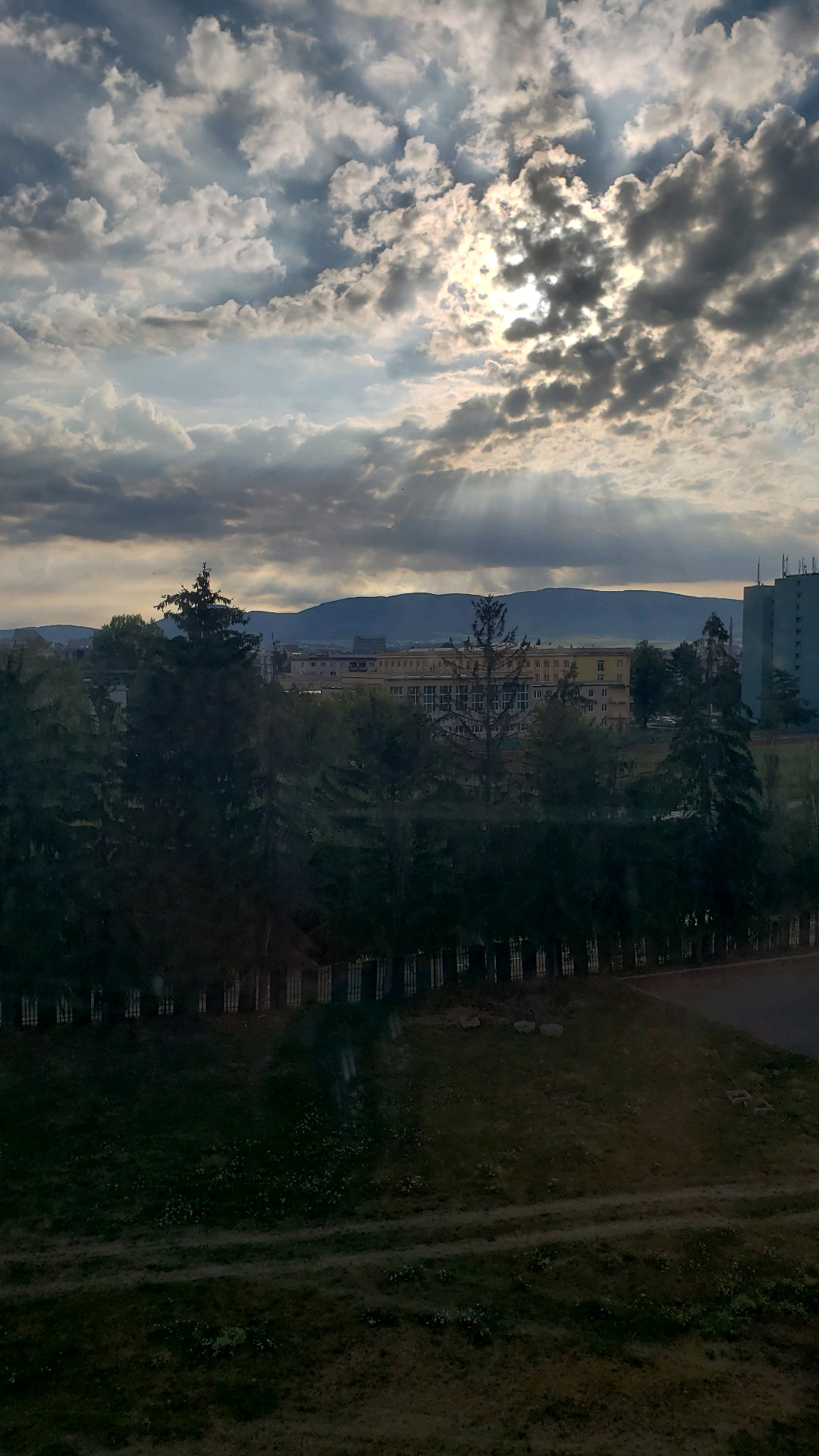 The image depicts a view of the city of Prešov, Slovakia, at dusk. The sky is filled with dramatic clouds, with shafts of sunlight breaking through, creating a crepuscular ray effect. In the mid-ground, a long, multi-story building with a yellow facade is visible, possibly an educational or institutional building, flanked by trees. To the right, a modern, tall, blue-tinted building stands out. The foreground consists of a grassy slope with a dirt path winding through it, and a dark, low fence runs along the edge of the vegetation. The overall impression is one of a somewhat overcast but visually striking evening sky over an urban landscape.