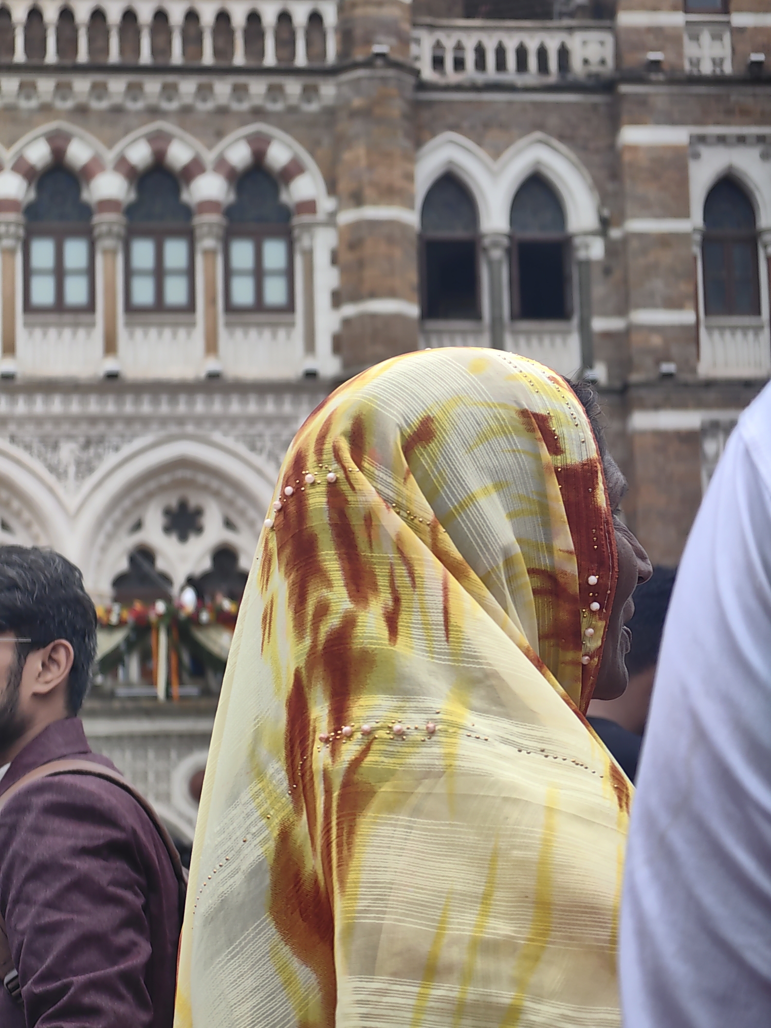 The image captures a close-up view of people in front of the Chhatrapati Shivaji Maharaj Terminus (CSMT) in Mumbai, India. In the foreground, a woman is partially visible, her head and shoulder covered by a yellow and red patterned dupatta adorned with small pearls. To the left, a man wearing a maroon jacket is also partially seen. In the background, the intricate architecture of CSMT is visible, with its arched windows and ornate detailing. The scene appears to be during daytime, with a slightly overcast sky. The overall impression is that of a bustling urban environment with a focus on the cultural and architectural elements of Mumbai.