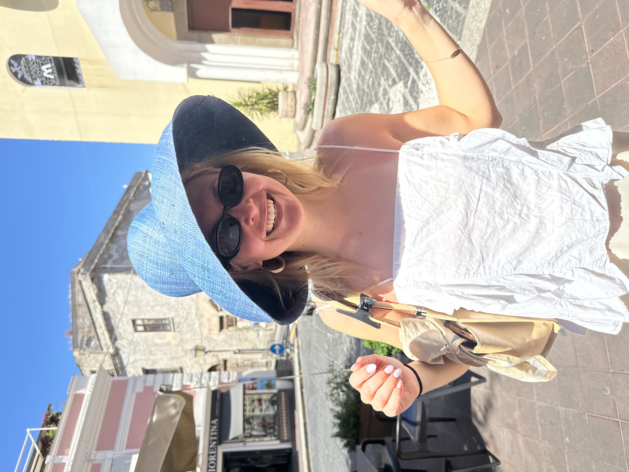 A vibrant outdoor portrait shows a smiling young woman against a sunlit street scene, likely in Lipari, Italy. The image is rotated 90 degrees counter-clockwise. The blonde-haired woman, centered in the frame, wears a light blue wide-brimmed hat with a dark band, and dark, round sunglasses. She is dressed in a white, crinkled top with spaghetti straps, displaying a wide, cheerful smile that shows her teeth. A gold hoop earring is visible on her left ear, and a delicate gold bracelet adorns her right arm. Her fingernails are painted a light, possibly white, color. She holds a light beige bag or garment carrier, which contains a mobile phone in one of its pockets.
The background features typical Mediterranean architecture: old buildings with textured walls in shades of yellow, off-white, and light pink, some with arched windows or doorways. The pavement is made of square stone tiles. A clear, bright blue sky dominates the upper portion of the frame, indicating a sunny, warm day, likely midday or early afternoon. Visible details include a "W" logo on an upper window and the text "FIORENTINA" on a shop sign. A blue circular street sign with a white arrow is also present. The woman's joyful expression suggests a relaxed and happy mood, possibly during a vacation or outing.