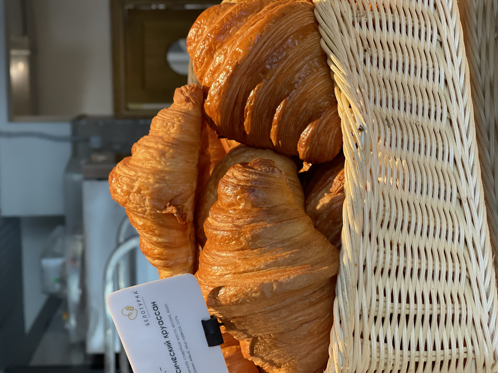 A basket of croissants is displayed in what appears to be a bakery. A white label with Cyrillic text is positioned in front of the croissants, partially obscuring them. The croissants are golden brown and flaky, stacked in a wicker basket. The background is out of focus, but appears to be an interior space, possibly a commercial kitchen or storefront. The lighting suggests indoor ambient light.