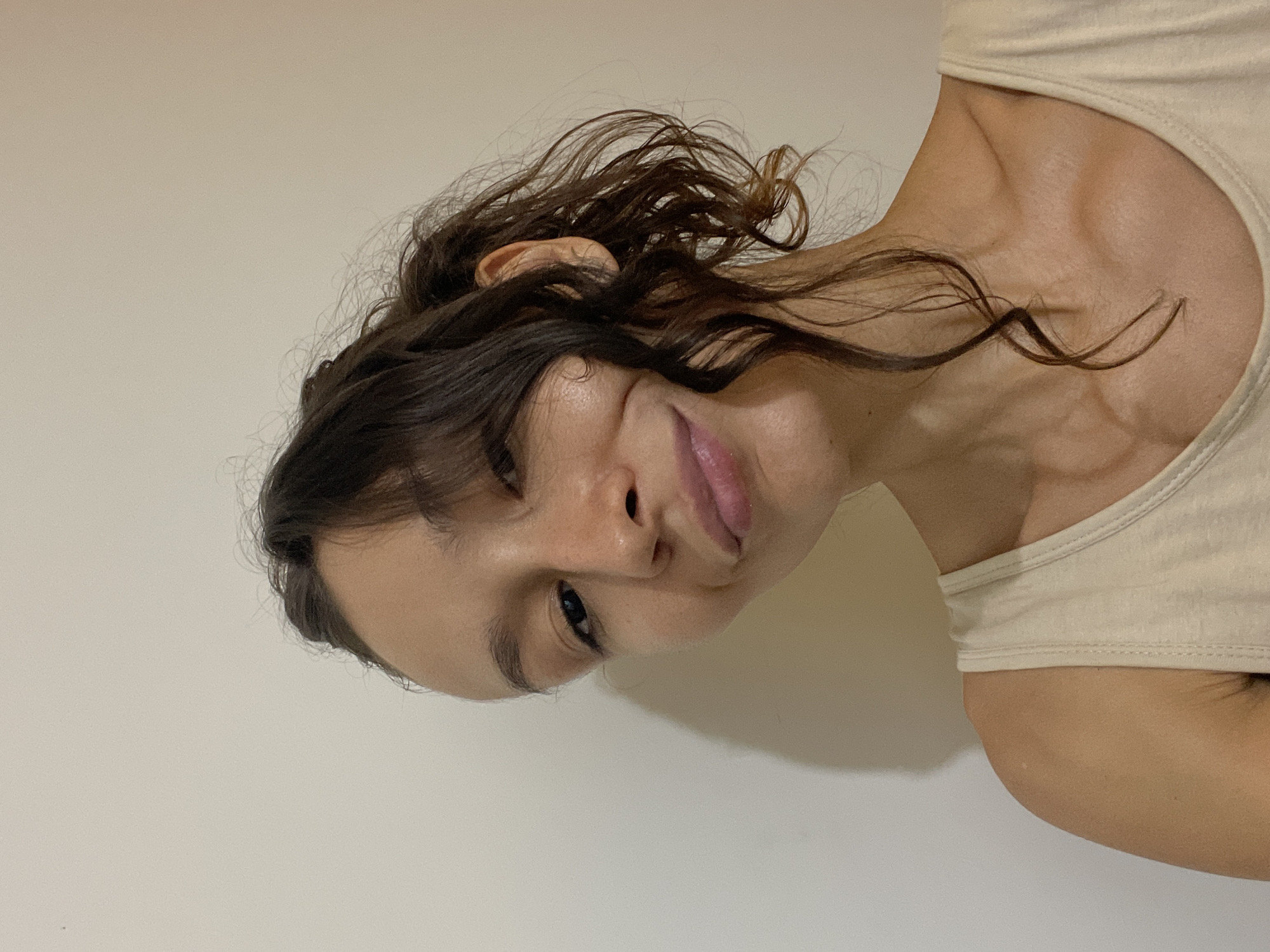 The media file presents an original image and several close-up crops, primarily featuring a candid portrait of a person, presumably a woman. The main image is rotated 90 degrees counter-clockwise, with the subject's head positioned on the left side of the frame, tilted as she looks directly towards the viewer.
The person has dark, wavy hair, which appears somewhat disheveled with loose strands framing her face and neck. A portion of her ear is visible through her hair. She is wearing a light-colored, possibly beige or off-white, tank top or camisole, revealing prominent collarbones and a slender neck. Her facial expression is gentle and relaxed, with one eye clearly visible looking at the camera. Her lips are slightly parted in a subtle smile or smirk, highlighted by a glossy pink shade. Her skin appears natural and smooth.
The background across all images is a plain, light-colored wall, indicating an indoor setting with no specific environmental cues beyond its simplicity. There are no other discernible objects or visible text in any of the frames. The close-up crops emphasize details such as the texture of her hair, the contours of her neck and shoulder, and the nuances of her facial features.
The overall impression is that of a casual portrait or selfie, captured in what appears to be natural or well-distributed artificial light, suggesting a daytime shot. While the image itself contains no specific location cues, the provided context places this scene in Mũi Né, Vietnam.