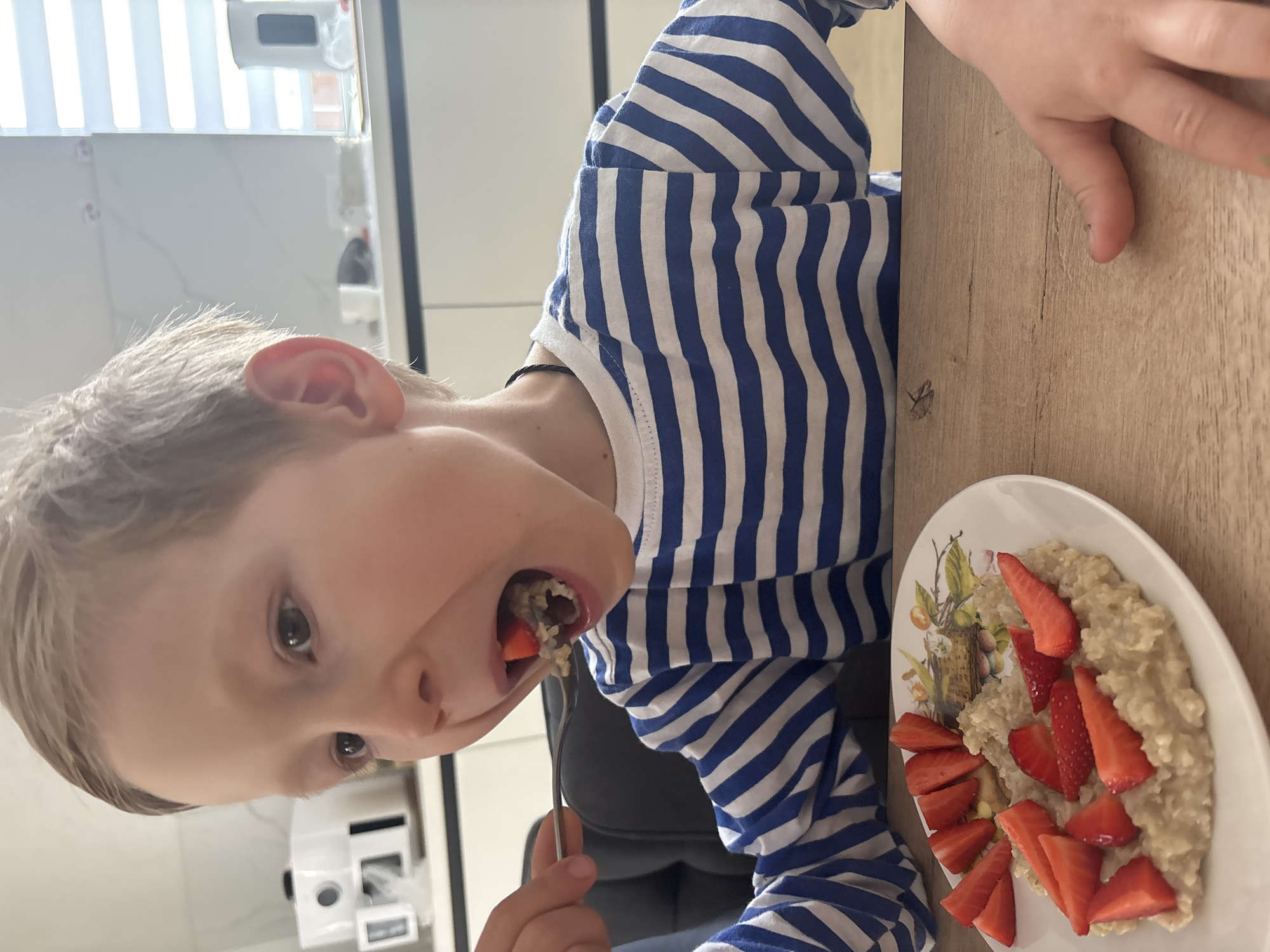A child, wearing a blue and white striped long-sleeved shirt, is eating oatmeal and sliced strawberries from a white plate. The child is holding a fork, with a piece of food partially in their mouth. The scene appears to be indoors, at a wooden table. In the background, there are white kitchen cabinets and blinds. The location is not provided.