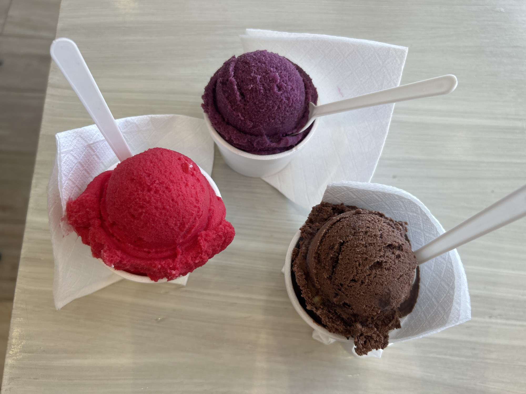 The image shows three small paper cups of ice cream, each with a small white plastic spoon. The ice cream flavors are red, purple, and chocolate. Each cup is wrapped with a white paper napkin. The cups are arranged in a slightly triangular pattern on a light-colored surface, possibly a table. The location is Unknown, Unknown.