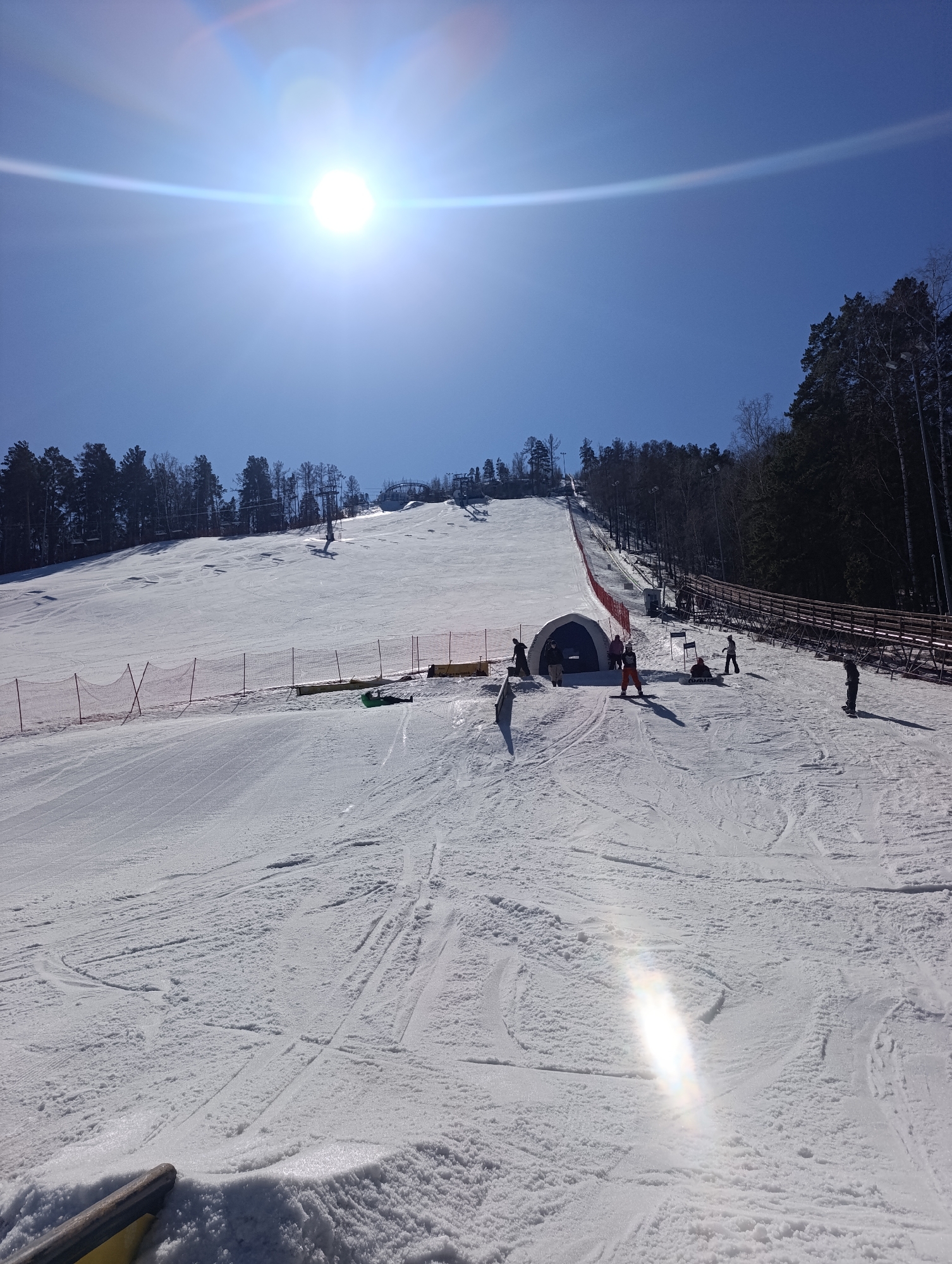 The image shows a sunny day at Bobrovy Log Funpark in Krasnoyarsk, Russia. A snow-covered ski slope is visible, with several people present. Some are standing near a dark tunnel-like structure, while others are skiing or snowboarding down the slope. Ski lifts are seen in the background, and trees border the edge of the slope. The sun is high in the sky, creating a bright glare. A wooden fence runs along the right side of the slope.