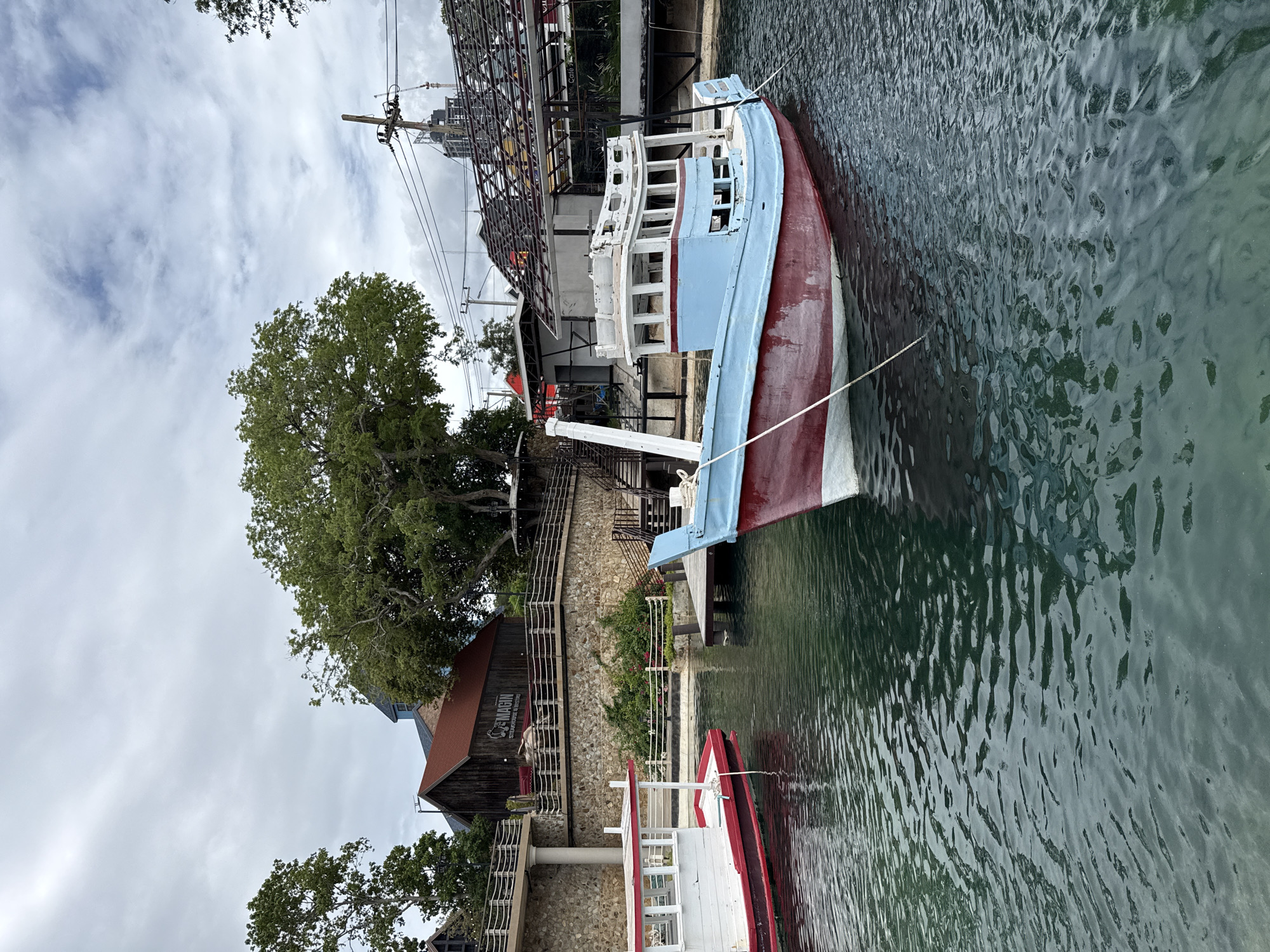 This is a photograph taken in Pattaya City, Thailand, depicting a waterfront scene with two boats docked in the water. The foreground shows the rippling surface of dark, green-tinged water. To the right, the bow of a blue and red fishing boat is partially visible, secured by a white rope to an unseen mooring. In the middle ground, a larger fishing boat with a light blue upper deck and a red hull is moored. A white rope connects it to a wooden jetty that extends into the water on the right side of the frame. To the left, another boat, predominantly white with a red stripe along its waterline, is also docked.
Behind the boats, a stone wall separates the waterfront from a raised embankment. On this embankment, a rustic, wooden building with a red roof stands, featuring the signage "MAGIN STEAKHOUSE & BISTRO". A set of stairs leads up to this building from the waterfront. More structures, including what appears to be a covered outdoor seating area and a building under construction with exposed metal framing, are visible further back. Large trees with lush green foliage frame the scene, and an overcast sky with scattered clouds covers the upper portion of the image. Electrical wires and poles are also present in the background, indicating an urban or semi-urban setting. There are no people visible in the image.