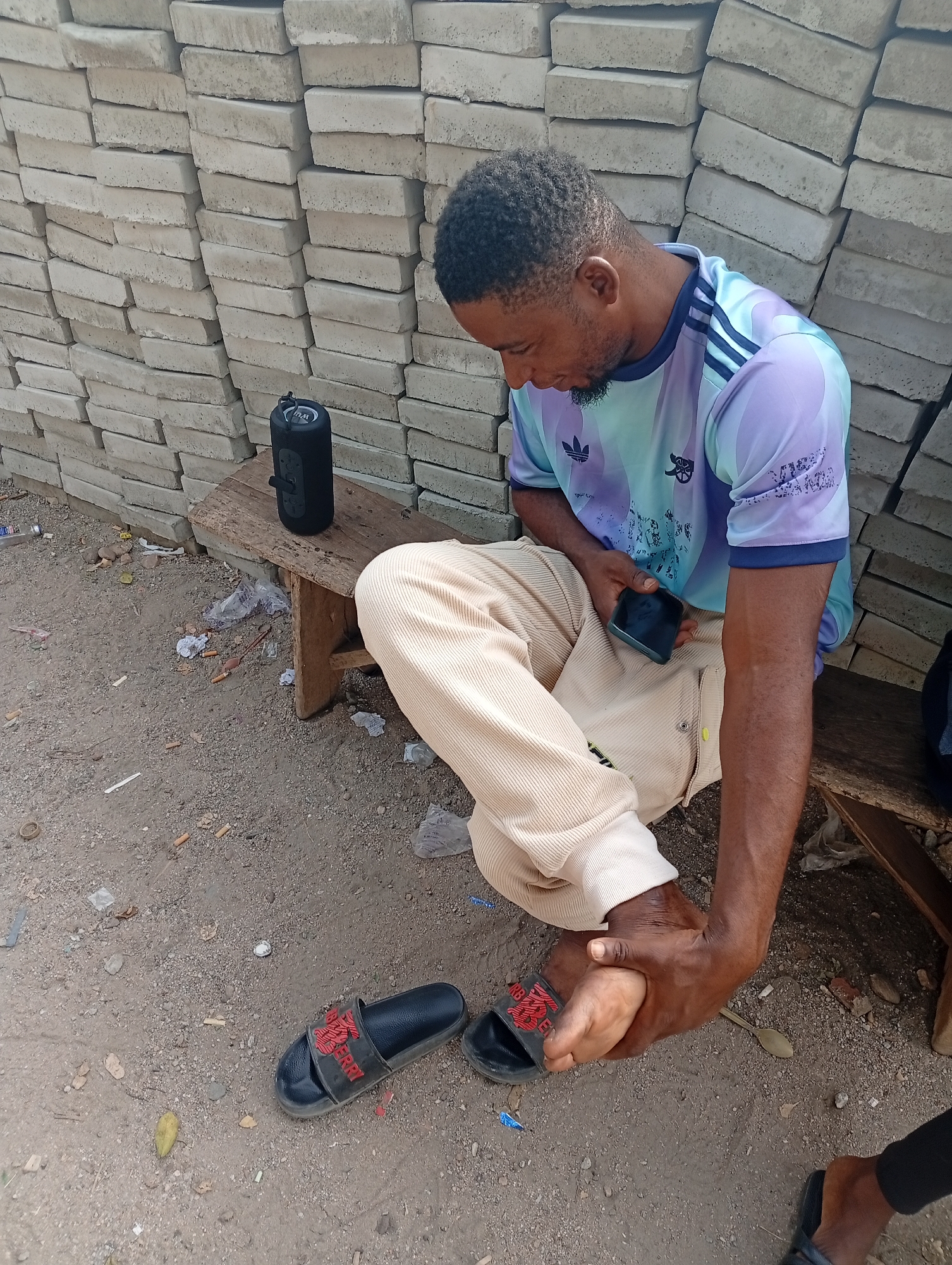 A man is sitting on a wooden bench outside in Adankolo, Nigeria. He is wearing a light blue t-shirt with dark blue stripes on the shoulder and light beige pants. He is holding a black cell phone in his left hand and holding his foot in his right hand, as if in pain. He has black slides next to him. Next to him on the bench is a black bluetooth speaker. Behind the bench is a wall of concrete pavers. The ground is dirt.