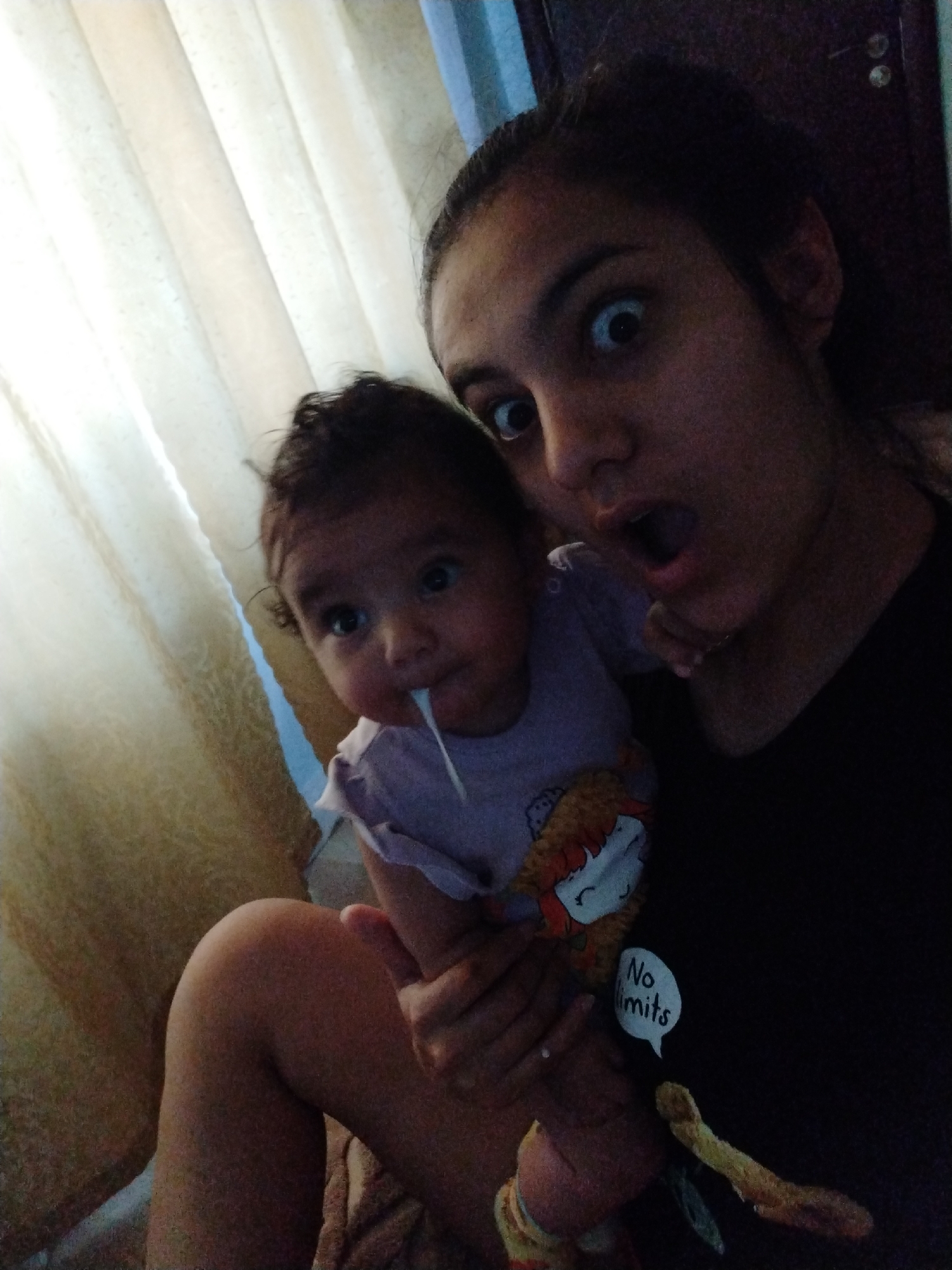 A young woman and a baby are in the foreground of a dimly lit indoor scene. The woman is taking a selfie, her face is close to the camera with wide, surprised eyes and an open mouth. The baby is nestled in her arms, with its head turned towards the camera. The baby has a string of drool coming from its mouth. The background shows a cream-colored curtain and a dark wooden door. The woman is wearing a black t-shirt with a graphic design and text that reads "No limits." The baby is wearing a light purple garment. The overall impression is a candid and playful moment captured in a home environment.