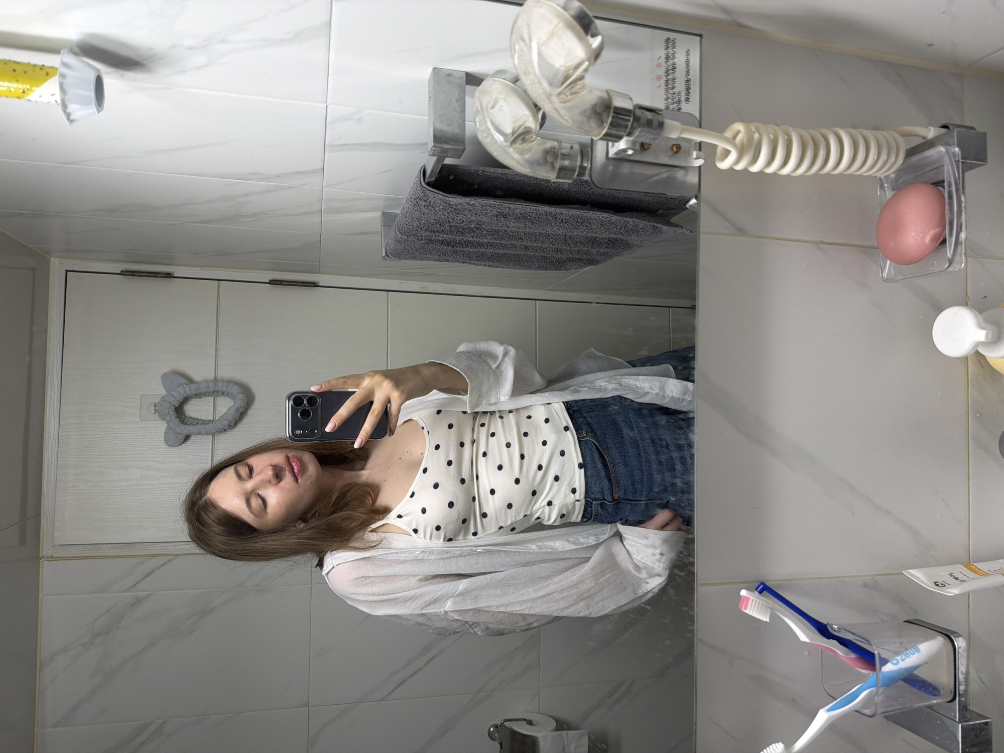 A young woman with brown hair takes a mirror selfie in a bathroom. She is wearing a white polka dot tank top and a white open button-up shirt, with denim shorts. She is holding her phone in her right hand. The bathroom has white marble patterned tiles on the walls and a white tiled floor. A towel rack with a gray towel is mounted on the wall. A handheld showerhead with a coiled white hose is also visible. In the foreground, two toothbrushes are in a clear cup, and a pink bar of soap is in a clear soap dish. The overall setting appears to be a residential bathroom in Dongducheon-si, South Korea.