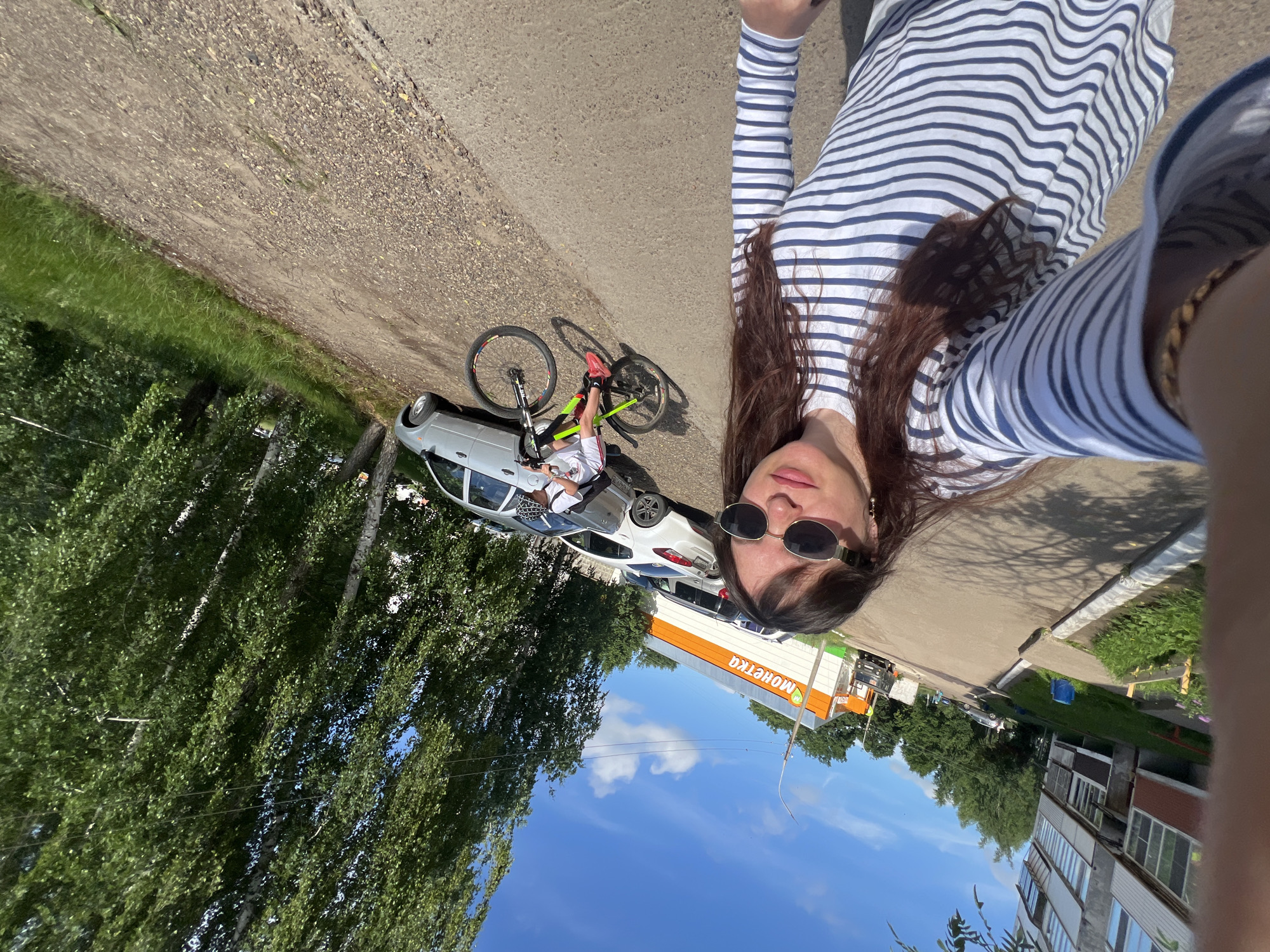 In Noviy, Russia, a woman wearing a striped shirt and sunglasses takes a selfie outdoors on a sunny day. Behind her, a child rides a bicycle on a dirt road. The background shows trees, cars, and a building with an orange and white sign that reads "МОНЕТКА".