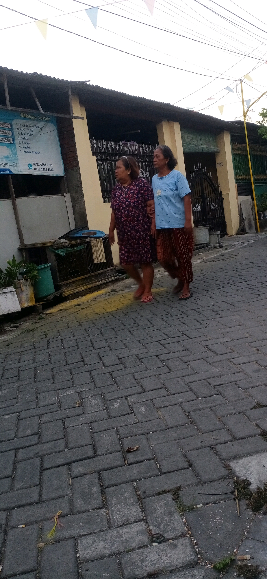 Two middle-aged women are walking arm-in-arm down a paved street in Sidojangkung, Indonesia. The woman on the left wears a dark floral print dress and pink flip-flops. The woman on the right wears a light blue t-shirt with a floral design and a patterned skirt. They are walking past a building with a yellow facade and a black wrought-iron fence. Bunting is visible strung across the street overhead. The street is paved with interlocking gray bricks. To the left of the building, a sign lists food items and phone numbers. Various potted plants and containers are visible near the building's base.