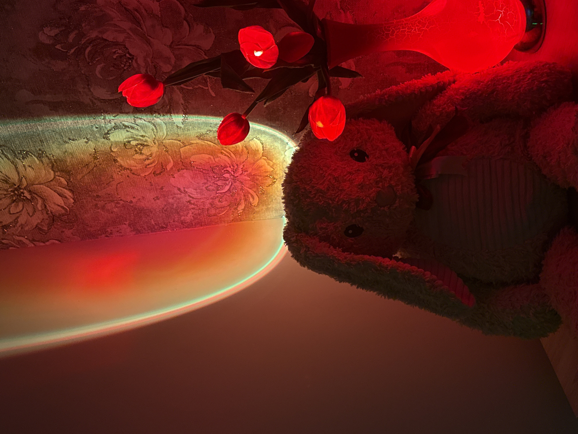 An indoor scene captured in dim lighting features a plush rabbit toy in the foreground, positioned to the left. Behind the toy, a wall displays a floral wallpaper pattern. To the right of the rabbit, a vase holds several red tulips. A bright, colorful light projection, appearing as a vertical band of red, orange, and green, illuminates the scene from the upper left, casting shadows and highlighting the subjects. The overall ambiance is warm and colorful due to the light's presence.
