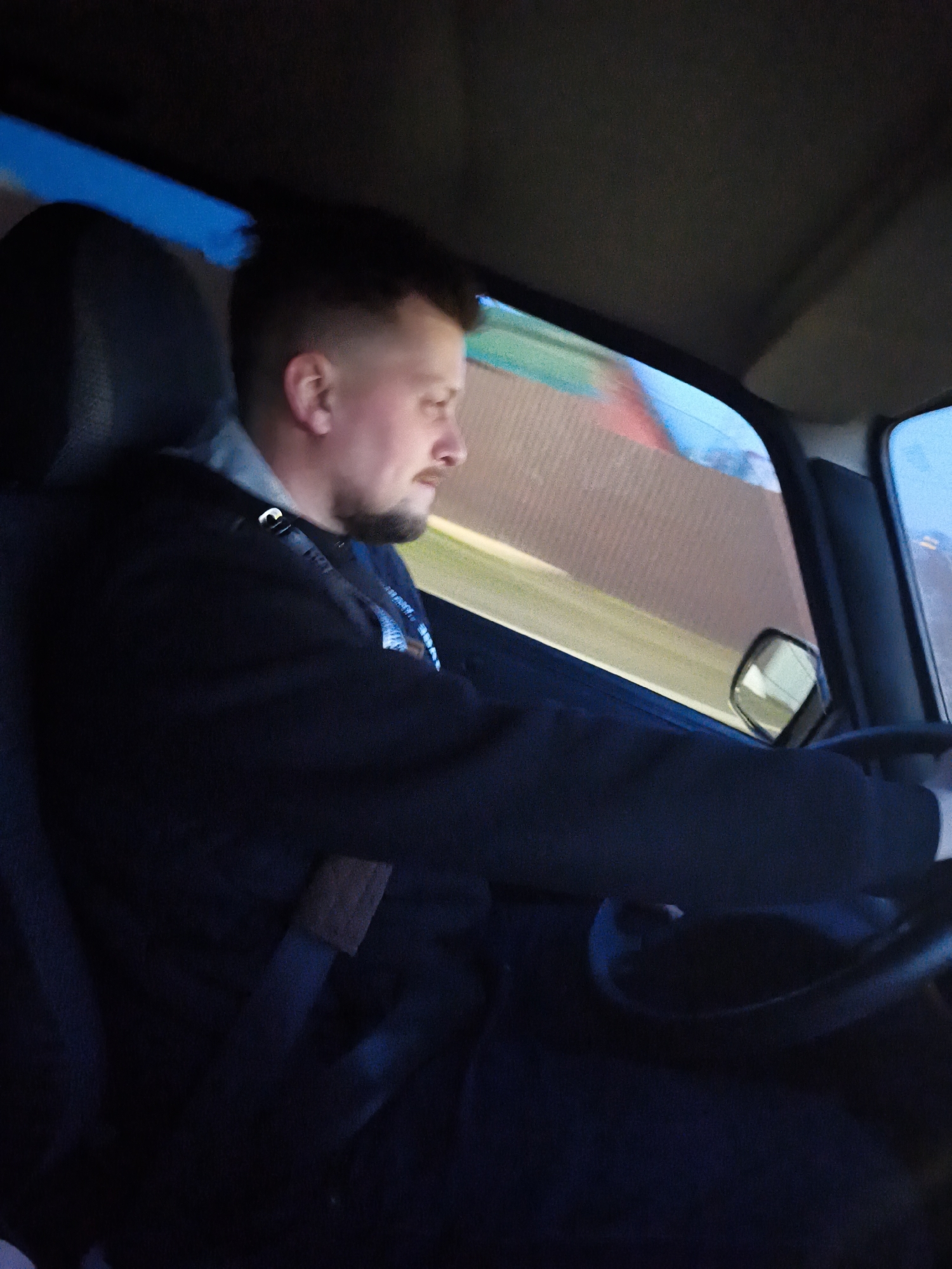 A man is driving a car, presumably in the city of Chaikovsky, Russia. The man is wearing a dark jacket with a hood and a seatbelt. The side view mirror is visible. Outside the window, a blurred fence or wall can be seen.