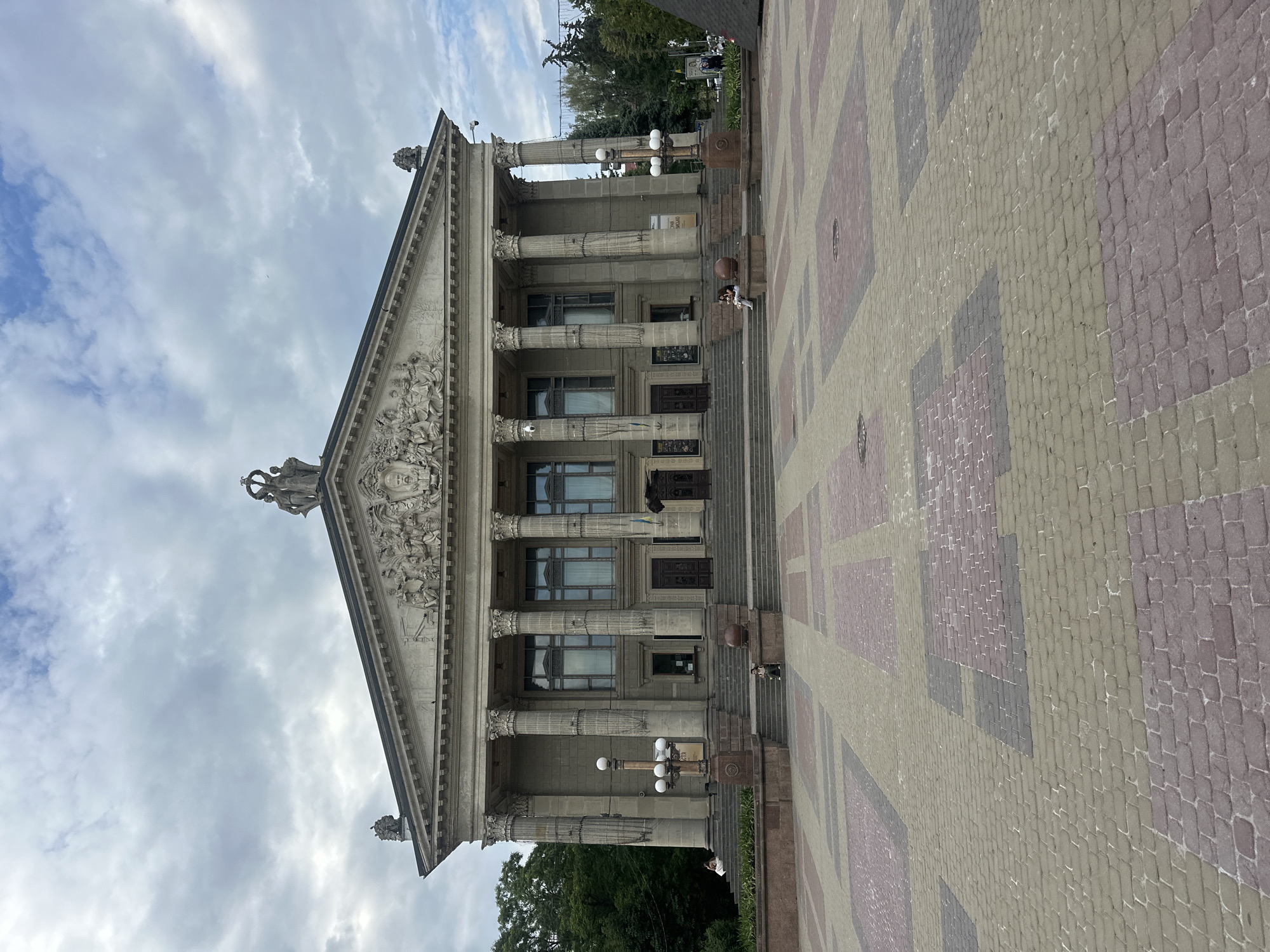 The image depicts the Ternopil Regional Drama Theatre in Ternopil, Ukraine. The grand neoclassical building features a prominent portico with six large columns supporting a pediment adorned with sculptures. A wide set of stairs leads up to the main entrance. The foreground is a pedestrian square paved with a geometric pattern of colored bricks. Two lampposts with multiple globes stand on either side of the stairs. Several individuals are visible on the steps and in the square, some seated and others walking. The sky is overcast with clouds. Trees and other buildings are partially visible in the background.