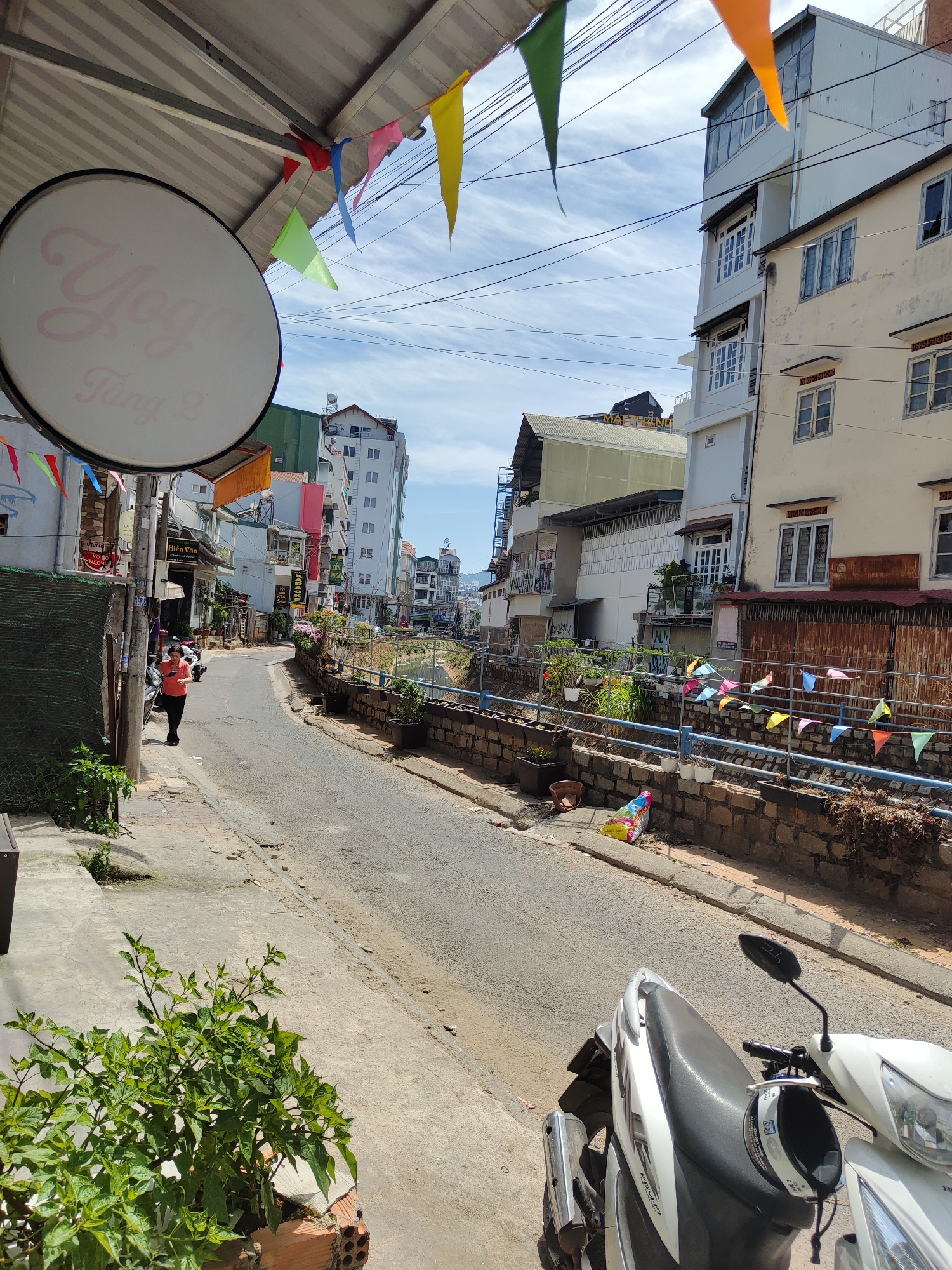 The image captures a street scene in Da Lat, Vietnam, with a narrow paved road winding through the urban landscape. Buildings of varying heights and architectural styles line the street, and a narrow waterway or channel runs along one side, bordered by a low stone wall decorated with small colorful flags. Plant life is present both in pots along the waterway and in a garden area on the left side of the frame. A white scooter is parked in the foreground, partially obscuring the view. A person is seen walking along the sidewalk on the left. Banners and flags are strung overhead, adding a festive atmosphere. The sky is partly cloudy, with hints of blue visible. A round sign with the text "Yoga Tang 2" is attached to a building on the left.