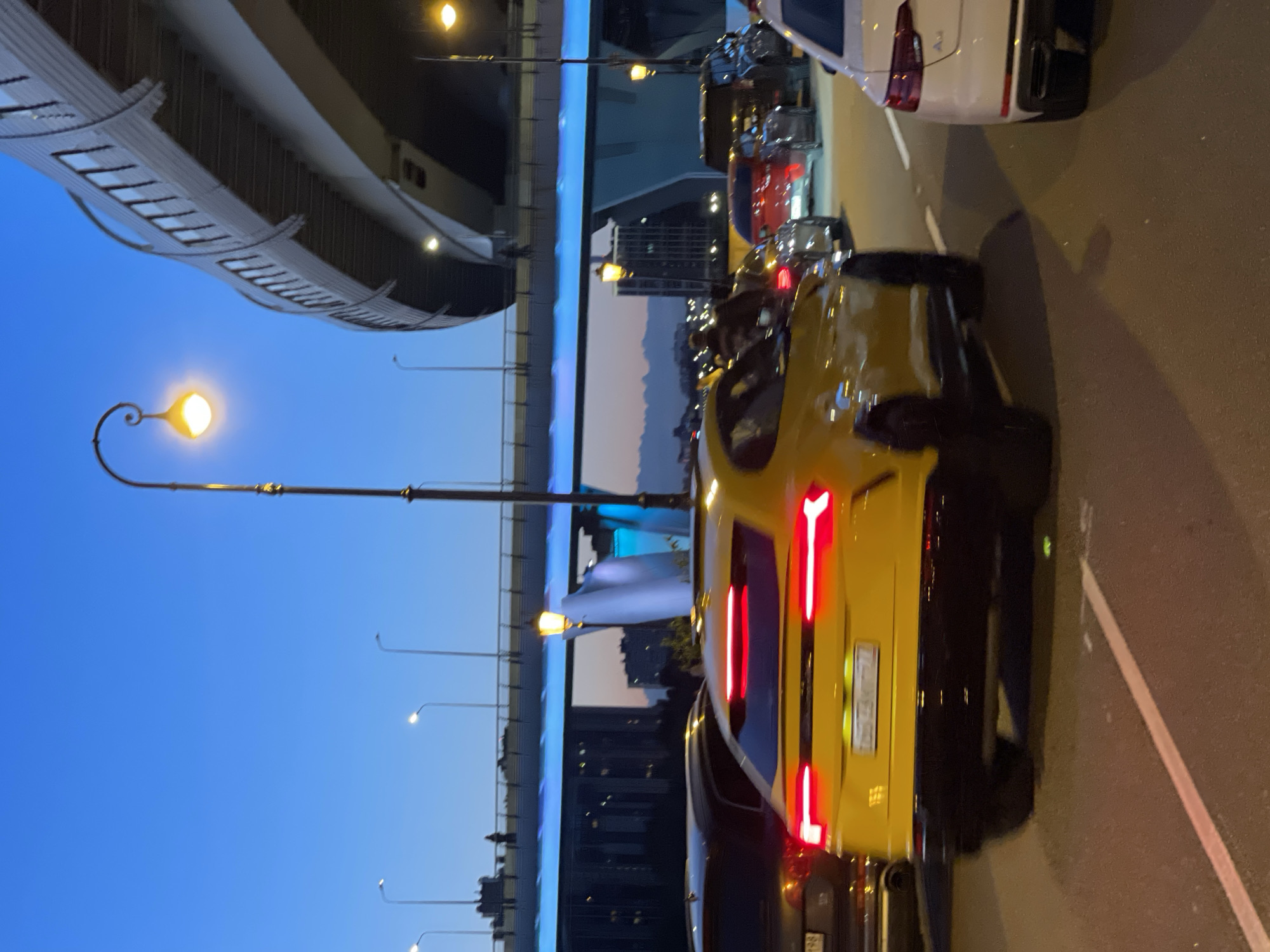 The image shows a street scene at dusk, likely in a city. In the foreground, a bright yellow SUV, possibly a Lamborghini Urus, is in motion, its brake lights illuminated. The car has a distinctive wide taillight bar. To its right, a white Audi sedan is partially visible. Other vehicles, including a dark SUV and a red car, are also in traffic. Overhead, a large, elevated highway structure dominates the background, with its lights illuminating the scene. Several streetlights with warm yellow bulbs are present, one prominently in the mid-ground with an ornate design. The sky is a deep blue, transitioning to a lighter hue near the horizon, suggesting twilight. Buildings and distant mountains can be seen in the background. The road surface is asphalt, marked with white lines.