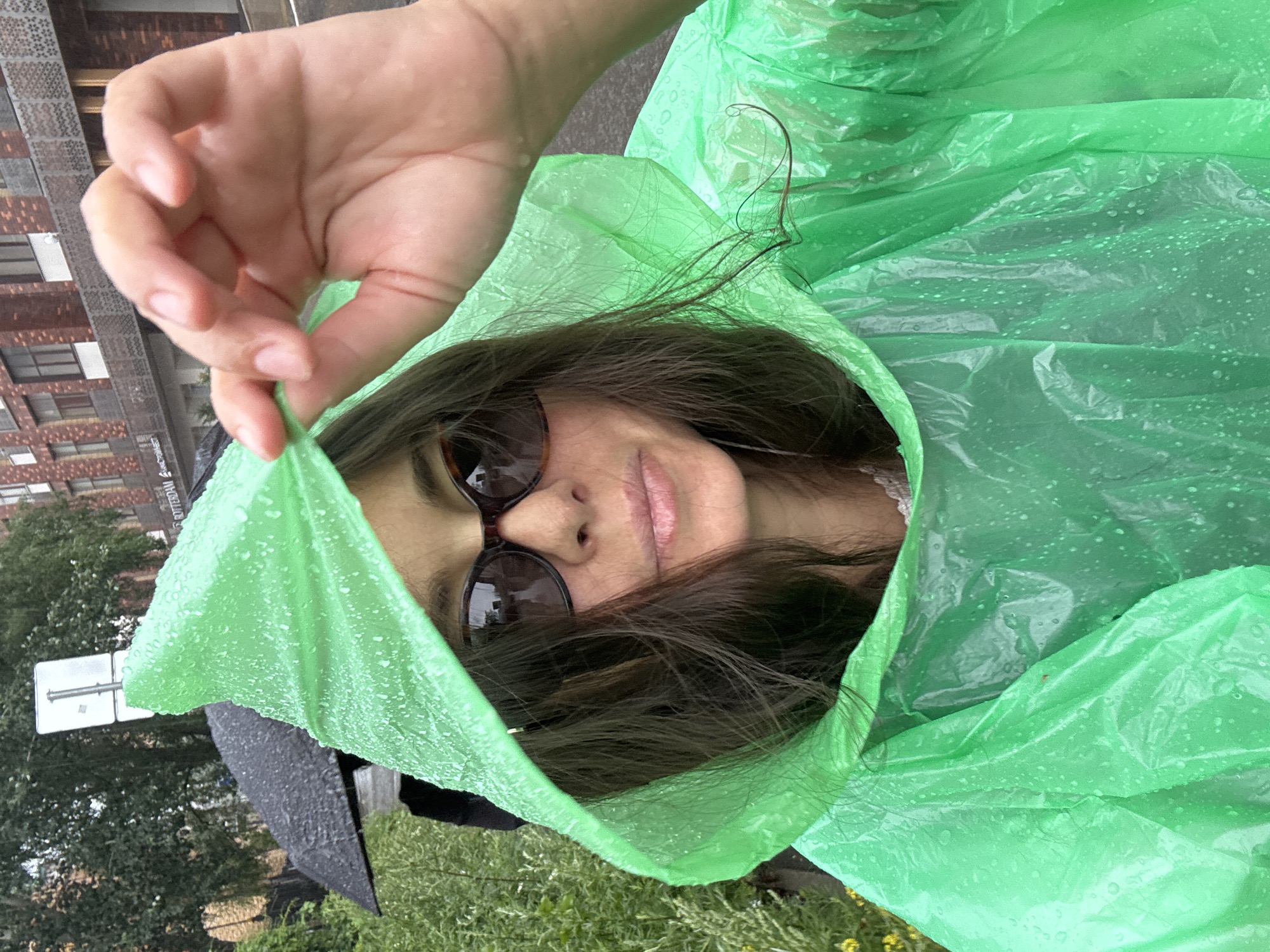 A person is captured in a close-up selfie, wearing a bright green, wet, hooded poncho. They are holding the edge of the hood open with their right hand, revealing their face. The person is wearing sunglasses and has dark hair. Their lips are slightly parted, and they appear to be smiling or smirking. The poncho is speckled with water droplets, indicating it is raining. The background is blurred, but shows greenery and the facade of a building. There's also a partially visible black umbrella. The overall impression is of someone braving the rain with a playful or adventurous attitude. The scene is outdoors, likely in an urban or suburban environment, given the building and street sign visible in the background. The time of day is not explicitly indicated, but the overcast sky suggested by the lighting implies it could be daytime. The location context is Kazan, Russia. No visible text is legible in the image.