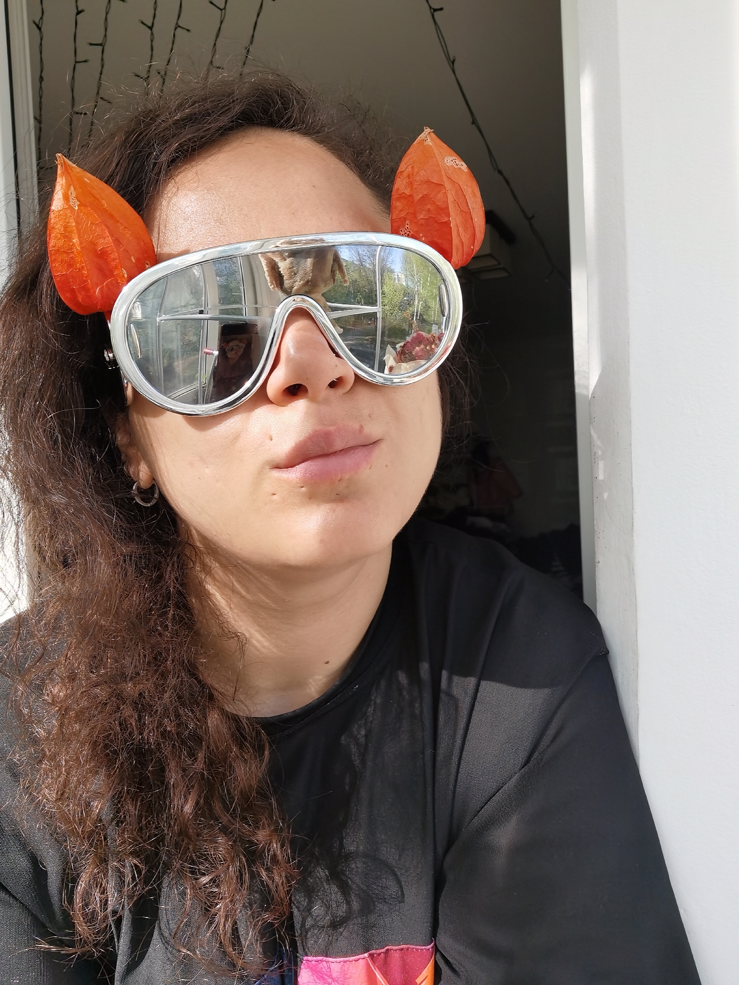 A woman is posing for a selfie in what appears to be a doorway or next to a wall. She is wearing a black t-shirt and silver framed glasses with orange physalis attached to the sides. She has curly brown hair, fair skin, and is wearing hoop earrings. Her lips are slightly pursed. The glasses reflect a scene of trees, sky, and what appears to be a person and a dog. Behind her, fairy lights are strung along the top of a window or doorway. The setting is likely indoors, near an open doorway or window. No information indicates this image's location as Chaikovsky, Russia.