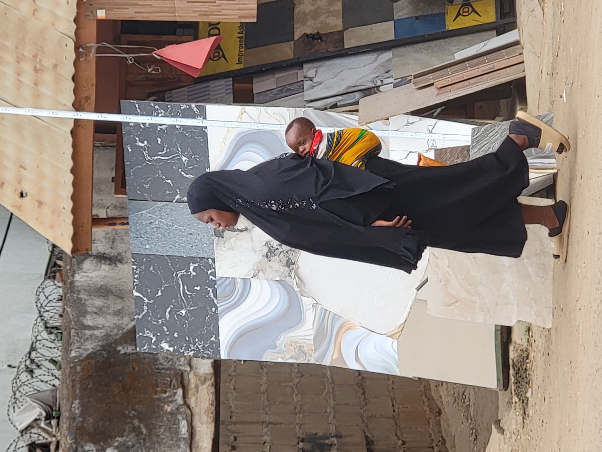 A woman wearing a black hijab carries a child on her back, standing amidst a display of ceramic tiles. The location is likely Maiburiji, Nigeria, suggested by contextual clues from the image, which include local architectural features, clothing styles, and potential product availability. The woman's attire suggests a Muslim cultural context. The backdrop features various tile designs, including marble patterns, likely part of a shop or street-side display. The ground is sandy and unpaved, hinting at an outdoor commercial environment.